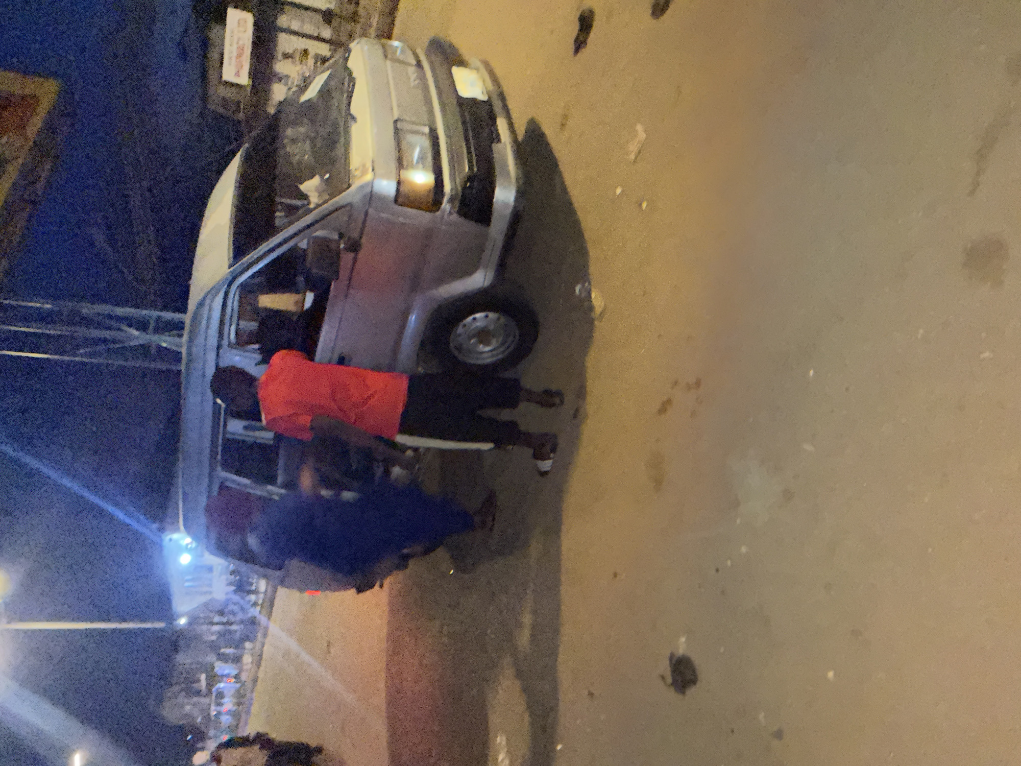 An over-the-shoulder, low-light, medium-angle shot captures a silver van parked on a paved street at night in Bukuru, Nigeria. Two individuals are visible near the van. One person, wearing a blue garment, appears to be standing by the rear passenger door of the van. The second individual, dressed in a red t-shirt and black shorts with white stripes, is leaning towards the driver's side of the van, appearing to interact with someone inside. The van's headlights are on, casting a glow on the street. The environment suggests a commercial or residential area, with some buildings and streetlights visible in the background. The ground is paved and appears somewhat dirty, with scattered debris.