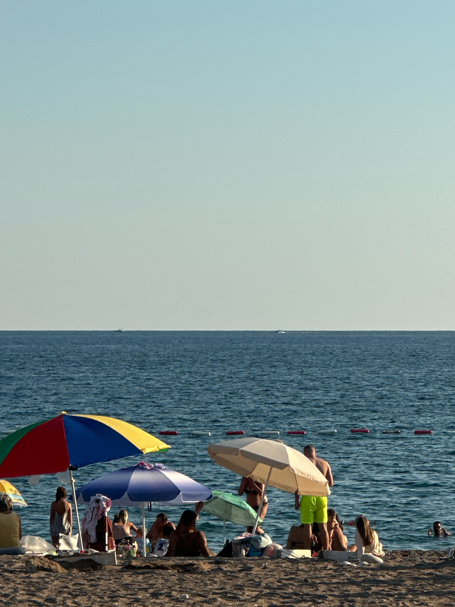A scene unfolds on a sun-drenched beach in Boreti, Montenegro. The foreground is a sandy expanse dotted with beachgoers seeking respite from the sun. Several umbrellas are strategically placed, offering shade. A vibrant, multi-colored umbrella with red, blue, and yellow segments stands on the left, partially obscuring a woman with dark hair and a patterned dress. To its right, a large, light-colored umbrella shelters a group of individuals. Among them, a man in bright green shorts stands, while others are seated or partially visible.
Further to the right, a green umbrella provides shade for another cluster of people, including a woman in a light-colored top and a man in the water, seemingly swimming. A pale blue umbrella is positioned between the multi-colored and the large light-colored one, with towels and bags placed underneath.
The middle ground is occupied by the calm, blue waters of the sea, stretching towards the horizon. In the distance, a small boat is visible on the vast expanse of water, and red and white buoys mark a section of the sea. The sky above is a clear, pale blue, transitioning to a slightly lighter hue near the horizon. The overall atmosphere suggests a peaceful day at the beach, with people enjoying the sun, sea, and sand.