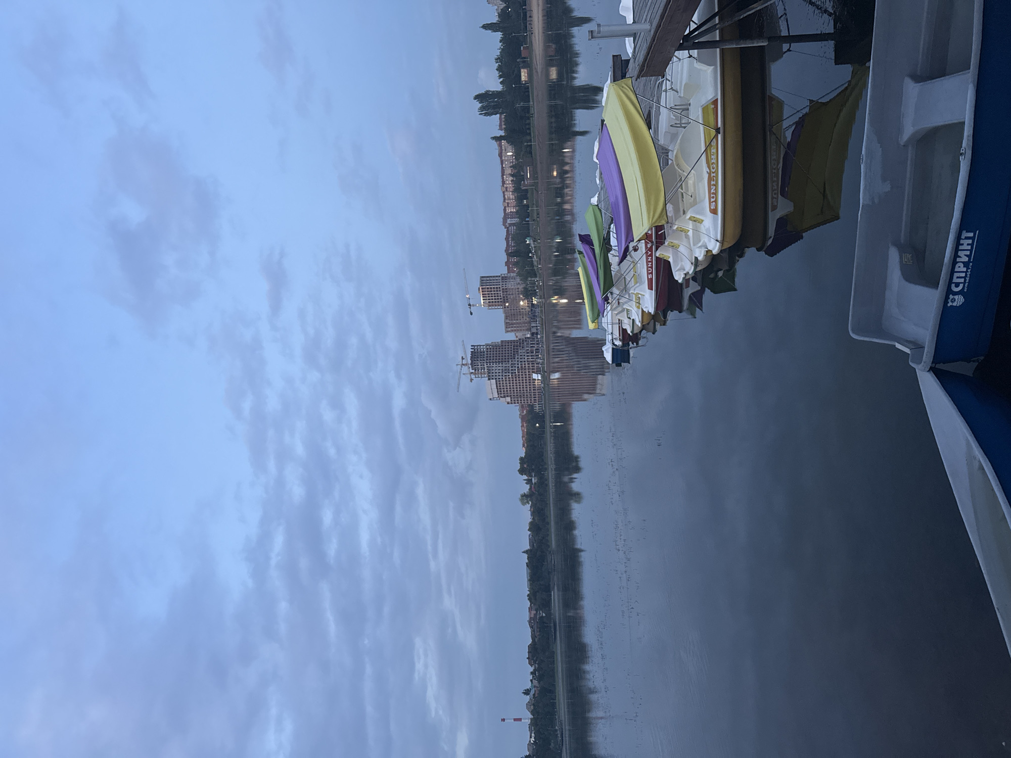 This image depicts a waterfront scene at dusk, likely on the Belaya River embankment in Ufa, Russia, given the presence of modern construction cranes and buildings characteristic of recent urban development in that area. The sky is overcast with diffused light, suggesting twilight or a heavily clouded day. In the foreground, several brightly colored paddle boats (yellow, purple, and green canopies) are moored to a wooden dock. These boats are designed for recreational use, with "SUNLIGHTS" visible on one. Further out on the calm water, which reflects the sky and distant shore, some aquatic vegetation pokes through the surface. The background shows a shoreline with a line of trees and buildings, including multi-story residential complexes and what appears to be a construction site with prominent cranes. A tall, red and white striped industrial structure, possibly a chimney or radio tower, is visible to the far left. The overall atmosphere is tranquil and slightly somber due to the low light conditions. No people are visible in the image.