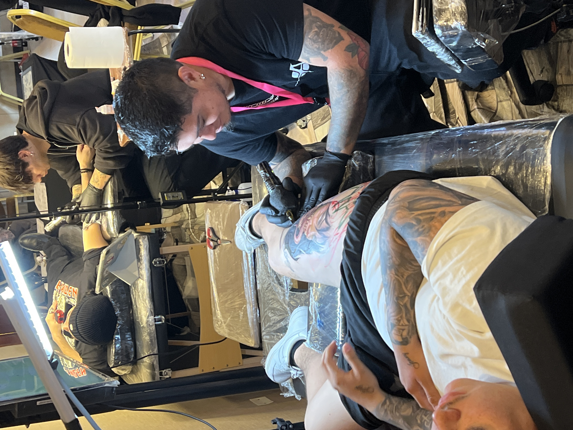 The image depicts a tattoo session in progress within what appears to be a studio or a convention setting, possibly located in the city of Arica, Chile. A tattoo artist, wearing black gloves and a black t-shirt with a pink lanyard, is actively working on a client's leg with a tattoo gun. The client, lying on a tattoo bed, has a partially completed, colorful tattoo on their thigh. The client is wearing black shorts and a white t-shirt. Another individual, also wearing black gloves and a black hooded sweatshirt, is seated and appears to be working on another client or supervising the session. The environment is characterized by various pieces of tattoo equipment, including a bright overhead lamp illuminating the workspace, a roll of paper towels on a stand, and what appears to be a tattoo chair or table covered in plastic wrap for hygiene. The background suggests an indoor setting with multiple tables and chairs, indicative of a busy event or studio.