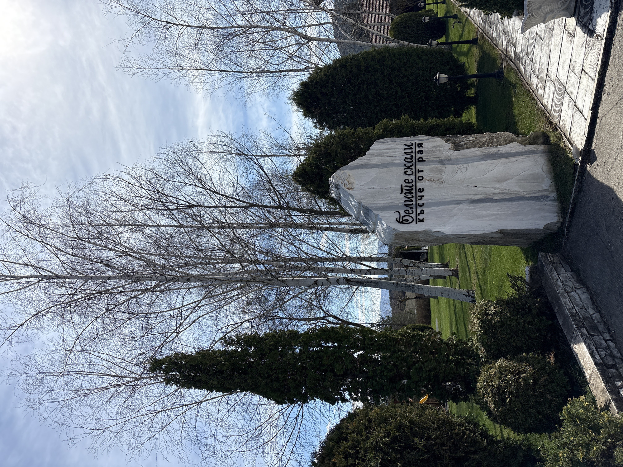 The image shows a stone monument with Cyrillic text in a park-like setting. The text appears to read "Белите скали кътче от рая". The monument is located near a paved pathway. The background includes deciduous trees without leaves and evergreen bushes. The sky is partially cloudy. The location is the city of Koprivlen, Bulgaria.