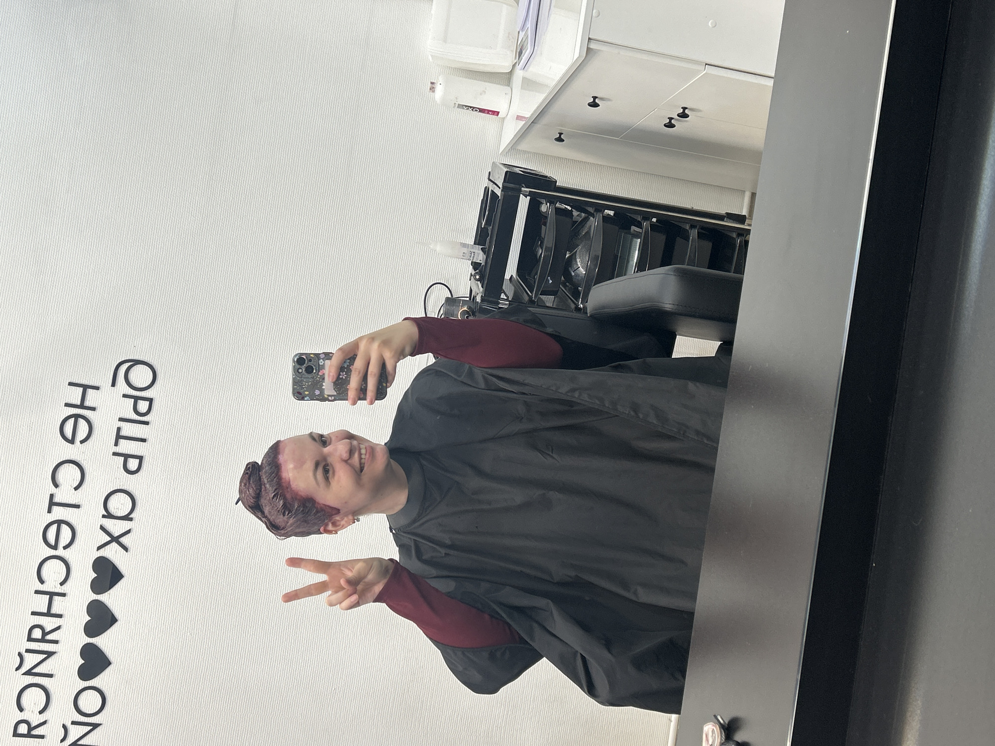 In Chaikovsky, Russia, a woman is captured in a mirror selfie within a hair salon. She smiles broadly, looking at her dark-colored smartphone, which has a floral-patterned case and is held in her right hand. Her left hand is raised, displaying a peace sign. Her hair, styled in a messy bun, is covered in reddish-purple hair dye, indicating a coloring treatment. She wears a maroon long-sleeved top beneath a black protective stylist's cape. The salon's environment features white, subtly textured walls. Above her reflection, mirrored black lettering on the wall reads "Я ЧУВСТВУЮ СЧАСТЬЕ НО ЛЮБЛЮ ВАС" (meaning "I FEEL HAPPINESS BUT I LOVE YOU"), interspersed with three black heart symbols. To her right, a black multi-tiered rolling cart holds various salon supplies, including bottles (one labeled "OXA") and bowls. Further to the right, a white cabinet unit with black pull handles supports additional white bottles and a small white digital scale. A dark, reflective countertop spans the immediate foreground.