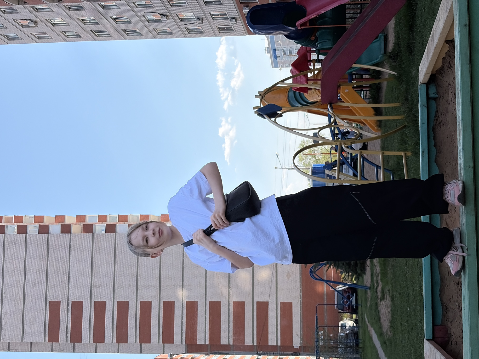 A vertically oriented shot shows a person and playground equipment against a backdrop of residential buildings and a clear sky.
The person, a child with blond hair, is positioned in the center of the frame wearing a white t-shirt and black pants. They are standing inside a playground's sandbox and holding a black purse.
Behind the child, colorful playground equipment is visible, including slides and climbing structures. The sandbox is constructed with green painted wood.
In the background, tall residential buildings with windows and brick-like designs are prominent on both the left and upper portions of the image. A clear blue sky is seen above.
Based on the available context, the location is likely in Izhevsk, Russia, though without further information, verification isn't possible.