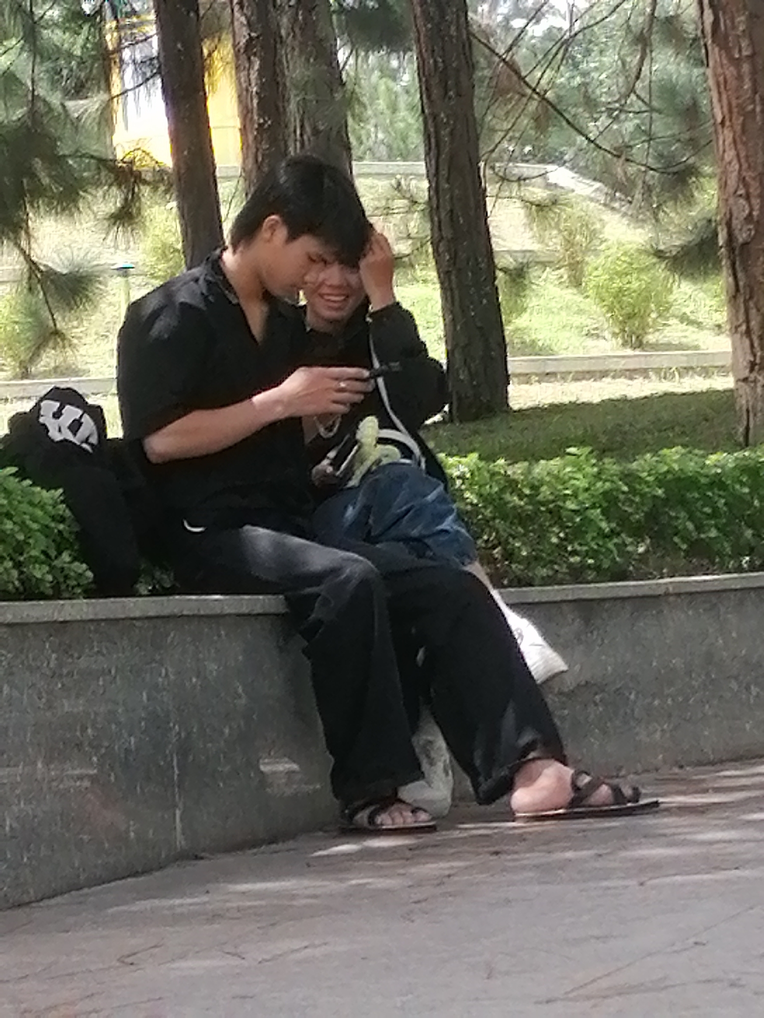 A man and a woman are sitting on a stone bench in a park, with trees and greenery in the background. The man is wearing a black shirt and black pants, and the woman is wearing a black jacket and blue jeans. They appear to be interacting with a mobile phone. The man is on the left, and the woman is on the right, leaning towards him. Their legs are crossed, and they are both wearing sandals. The man's left hand is holding the phone, and his right hand is touching the woman's head. The woman's left hand is resting on her lap, and her right hand is on the man's arm. The scene suggests a relaxed, intimate moment between the two individuals in a public park setting. The location is identified as Da Lat, Vietnam, a city known for its pine forests and temperate climate, which aligns with the visual cues of the image.
