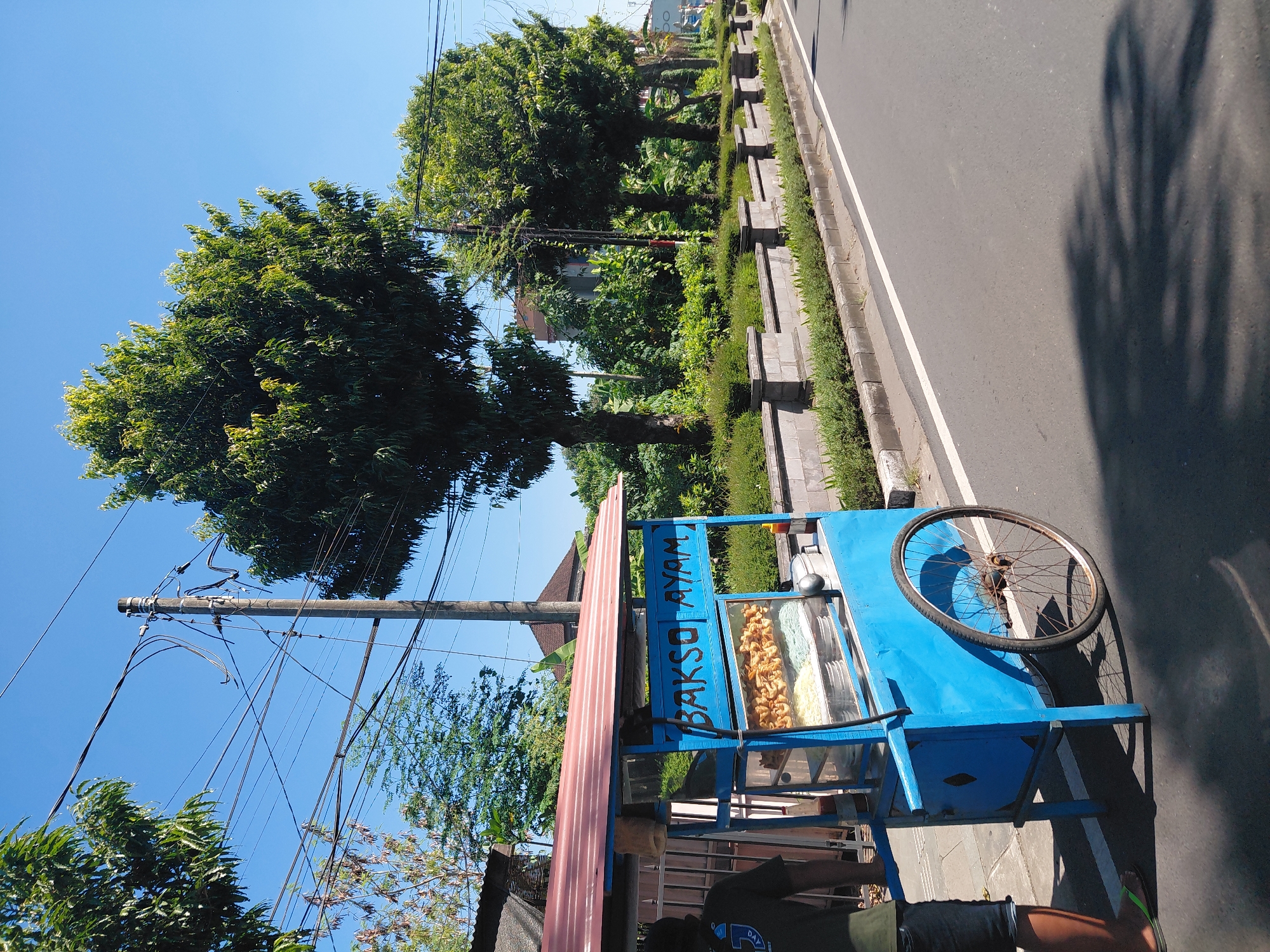 This image depicts a sunny roadside scene in Gianyar, Indonesia, featuring a blue and red "Bakso Ayam" (Chicken Meatball) street food cart. A person, whose lower legs and torso are visible, stands behind the cart on the left, wearing a dark shirt, dark shorts, and green flip-flops. The cart has a corrugated red roof and a glass display section showing various items: numerous golden-brown fried snacks on an upper shelf, and multiple metal bowls containing white and light blue noodles on a lower shelf. A small red bottle, likely a condiment, is on the right side of the cart's counter. The cart has a large, spoked wheel with a rusty hub on its right side.
The cart is positioned on a paved asphalt road, which features a solid white line marking its edge. To the right of the road, a concrete curb separates the pavement from a manicured planter area with a low stone wall and green shrubs. Numerous large, green-leafed trees line the road on both sides, with dense foliage obscuring buildings in the background. A tall utility pole with multiple crisscrossing black power lines and cables is prominent against a clear, bright blue sky in the upper-mid section of the image. Strong sunlight casts distinct shadows of the trees and the food cart onto the road surface.