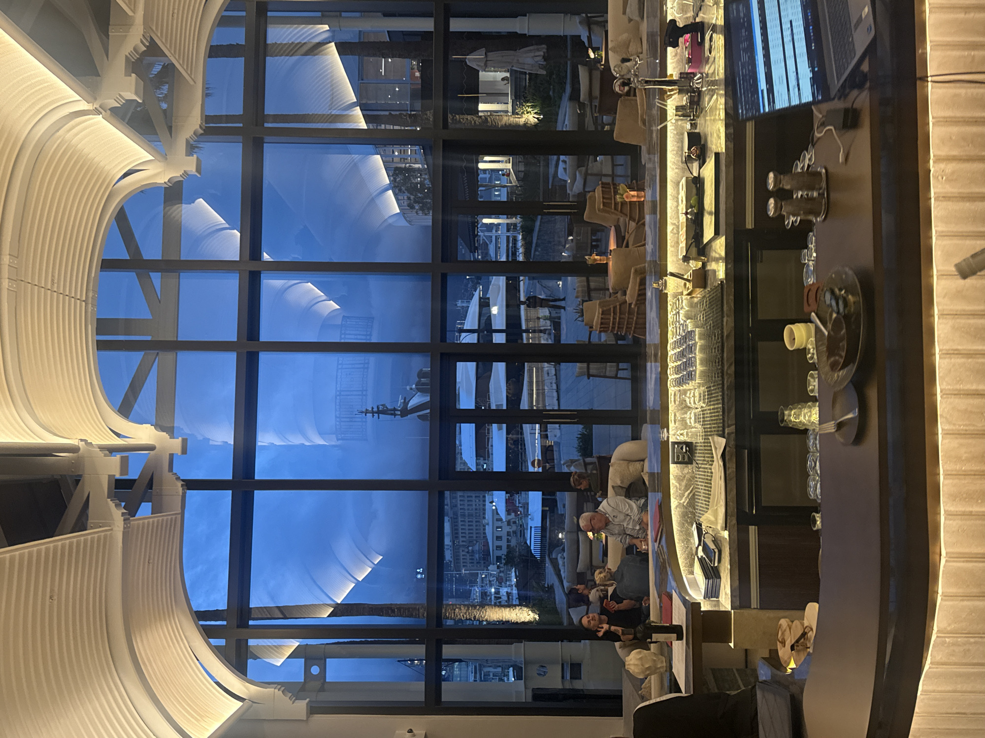 The image shows the interior of a bar or restaurant, likely in Cape Town, South Africa. Through a large, multi-paned window, a view of the city at dusk is visible. A harbor, cityscape, and other buildings can be seen in the reflection. Indoors, several people are sitting on couches and appear to be having a conversation, in the foreground right is the bar with many glasses and bottles. The bar is lighted and a computer display is visible at the bar. The building's ceiling is marked by a curved, white architectural feature. The photo is rotated 90 degrees clockwise, therefore some elements appear sideways or upside-down.