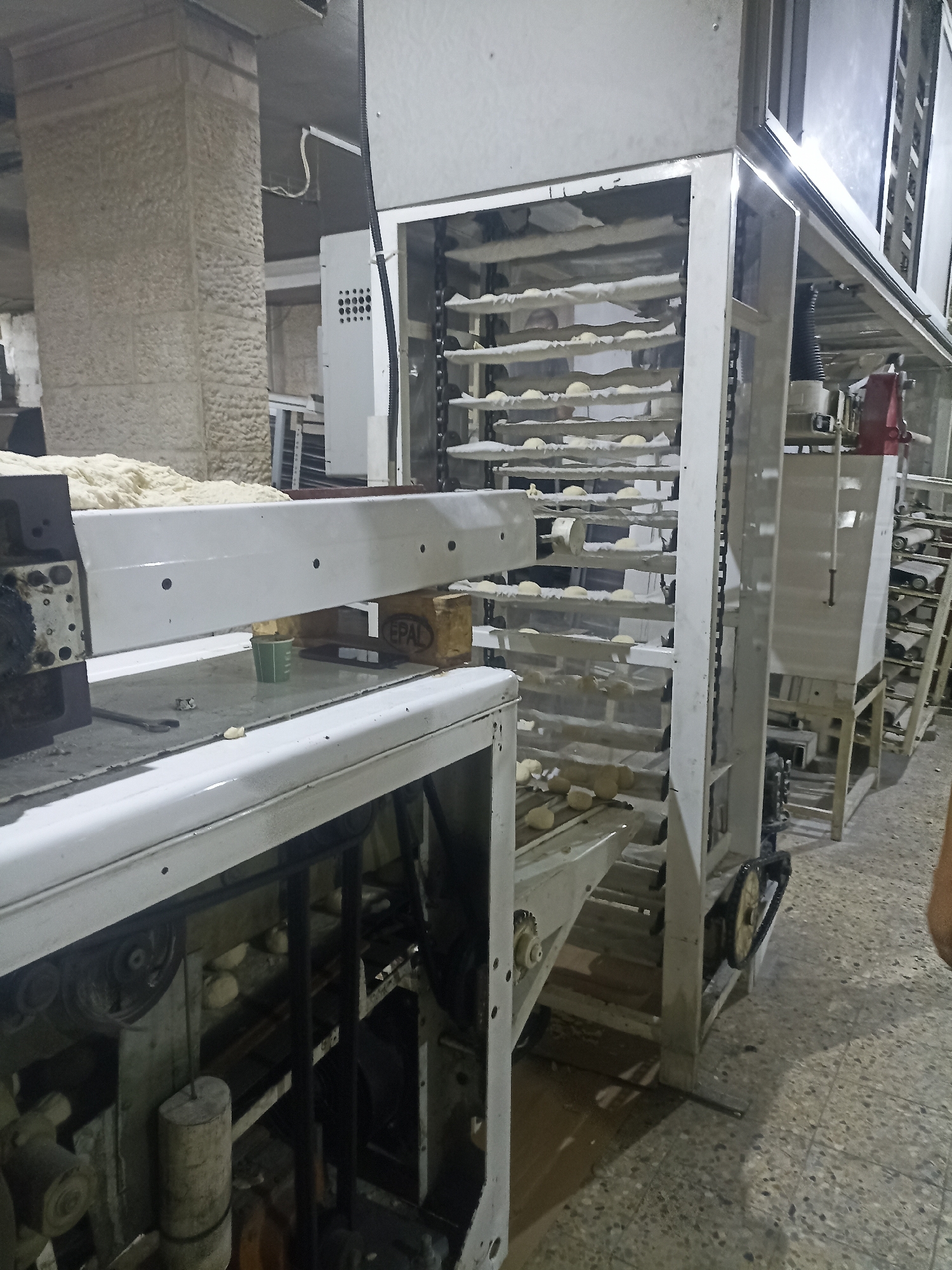 The image shows a view inside a bakery in Gaza City, Palestinian Territories. The scene is dominated by a large, multi-tiered bread-making machine, likely an oven or proofer. Several shelves are visible, laden with raw dough balls, some of which appear to be shaped into small loaves. The machine is constructed from metal, with visible chains, belts, and mechanical components. To the left, a conveyor belt system is partially visible, also carrying dough. In the background, a textured stone wall suggests the building's construction. The flooring is a speckled gray tile. The lighting appears to be artificial, common for an indoor industrial setting. There are no people clearly depicted in the frame, though their presence is implied by the operation of the bakery equipment.