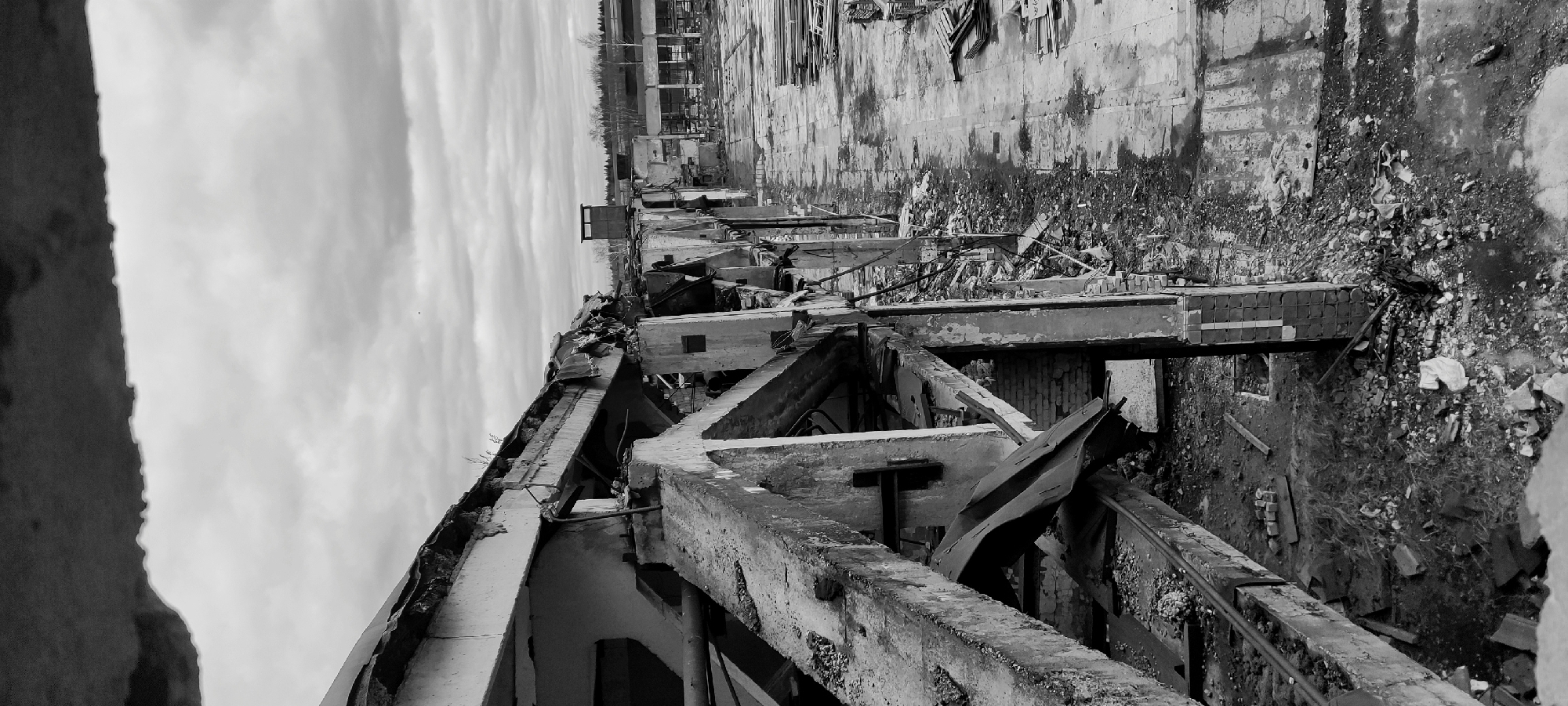The black and white image, presented in a portrait orientation but depicting a landscape scene, shows the extensive ruins of a large building or industrial complex. The scene is dominated by debris and damaged concrete structures under an overcast sky.
In the foreground, skeletal remains of concrete beams, pillars, and fragmented walls are visible, stretching from the lower left towards the upper right. These structures are heavily damaged, with rebar exposed and sections appearing collapsed or twisted. Various pieces of rubble, including bricks and metal, are scattered throughout this area.
The middle ground reveals more piles of debris and the foundations of what were once rooms or sections of the building. A distant, partially intact wall with signs of wear and damage forms a backdrop to the immediate destruction. Beyond this, in the far background, a line of trees and other distant, less damaged buildings can be discerned.
The sky, occupying the upper portion of the re-oriented image, is uniformly cloudy, suggesting an overcast day. No people are present, and there are no discernible activities occurring in the scene. The overall atmosphere conveyed is one of desolation, abandonment, and the aftermath of significant destruction. No visible text can be clearly read.
The context provided for the location is Fryazino, Russia.