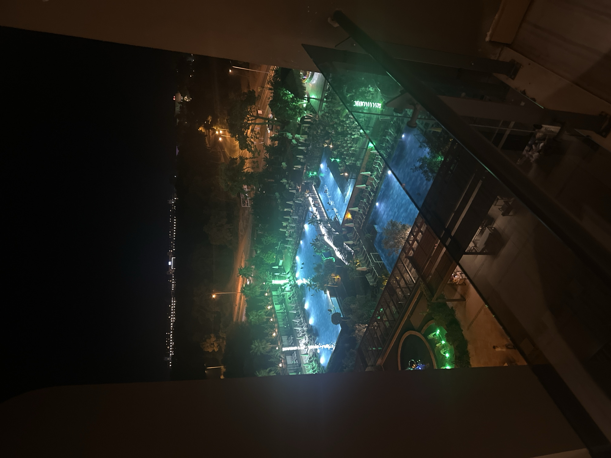 This is an aerial, nighttime photograph taken from an elevated vantage point, looking down upon an illuminated resort complex. The main focal point is a large, irregularly shaped swimming pool, brightly lit with blue underwater lights. Several individuals are visible swimming and relaxing in the pool. Surrounding the pool are numerous lounge chairs and umbrellas, many of which are also illuminated by green lighting. Beyond the pool area, there are palm trees and other vegetation, with more lights indicating pathways and architectural features. In the distance, a cityscape with numerous lights suggests the presence of buildings and infrastructure. The foreground of the image is dominated by the dark interior of a building or balcony, with a glass railing reflecting the green lights from the resort. The image appears to have been taken in Manavgat, Turkey, likely at a resort that utilizes extensive nighttime illumination to create a vibrant atmosphere.
