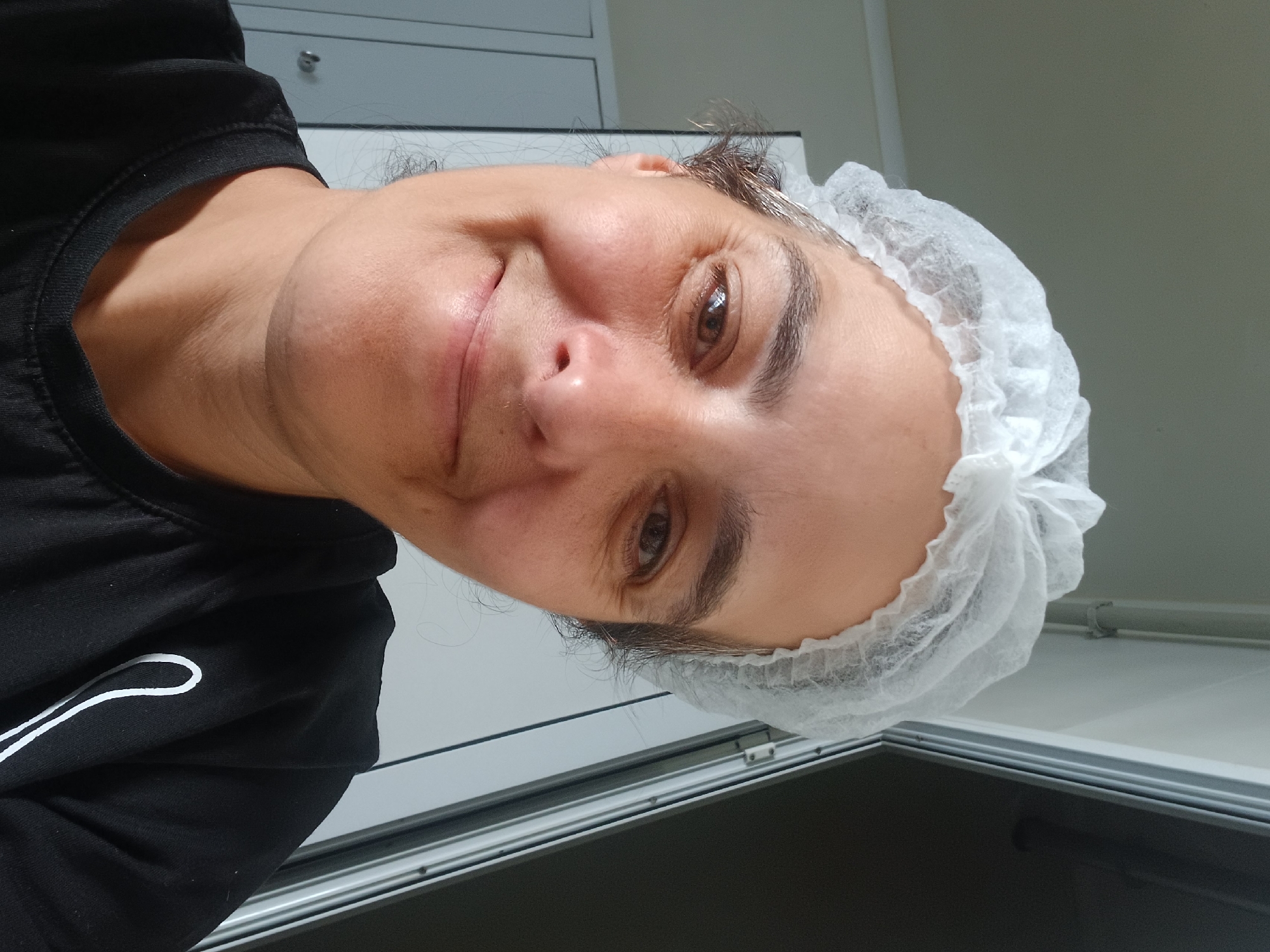 A person is shown in a close-up, sideways selfie, wearing a black t-shirt and a white disposable hair net. The person is smiling slightly at the camera, with their head tilted. Their eyes are brown and they have dark eyebrows. The background is blurred, but appears to be an indoor setting with neutral-colored walls and cabinets. The lighting suggests it is daytime. There is no visible text in the image. The context provided indicates the location is Rakevo, Bulgaria.