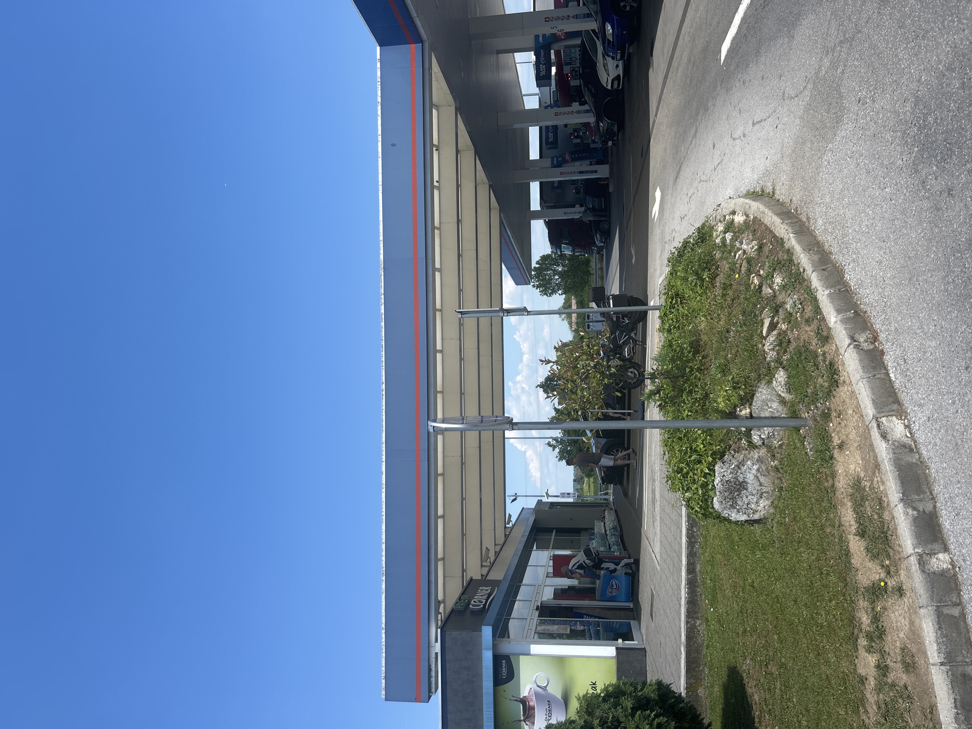 An outdoor medium shot shows a gas station on a clear, sunny day. The sky is a vibrant blue with a few wispy clouds. In the foreground, a gray asphalt road curves to the right. A concrete curb separates the road from a grassy area with scattered rocks and bushes. A silver metal pole stands in the grassy area. Beyond the grass is a sidewalk and the entrance to a store with large glass windows. A person in a blue shirt and black pants is standing in the doorway, looking towards the inside of the store. Several people are visible near vehicles under the gas station's canopy. A red semi-truck is parked at a pump, and a dark-colored car and a white car are also visible. A motorcycle is parked on the left side of the frame. The gas station canopy is supported by white pillars. The scene is illuminated by bright sunlight.