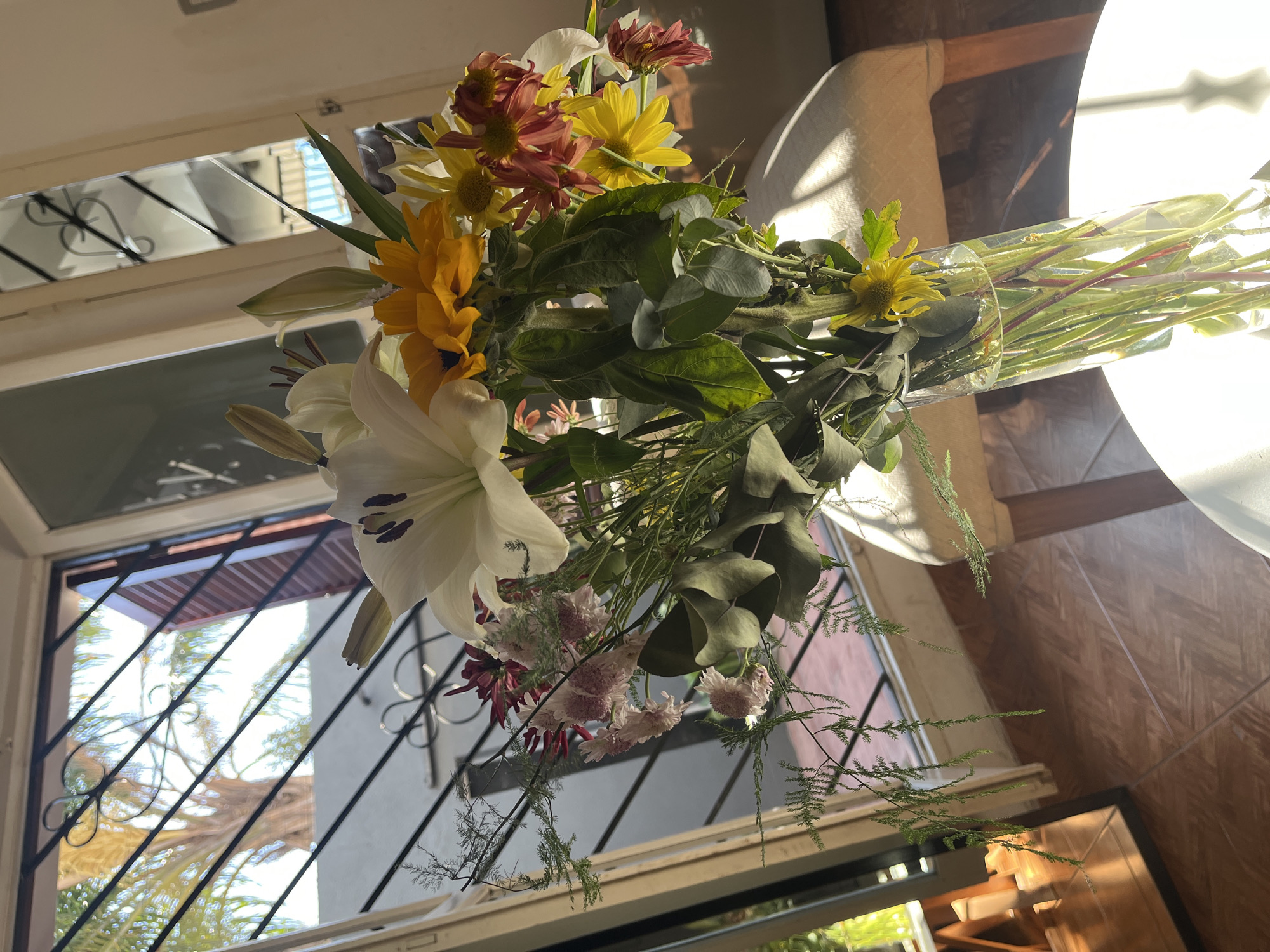 A bouquet of mixed flowers sits on a surface indoors, possibly a table or windowsill, with a window in the background. The flowers include a large white lily with dark purple stamens, yellow sunflowers, orange daisies, and smaller pink and white flowers. Green foliage is interspersed among the blooms. The bouquet appears to be in a clear vase or wrapping.
The window behind the flowers has a decorative metal grille and allows a view of trees and buildings outside. Based on the distinctive architectural style visible through the window, this could be an exterior view of San Fernando del Valle de Catamarca, Argentina. The indoor environment is bathed in natural light, suggesting a daytime setting. The walls visible behind the flowers appear to be wooden. A white object, possibly a lamp or part of the furniture, is partially visible to the right of the flowers.