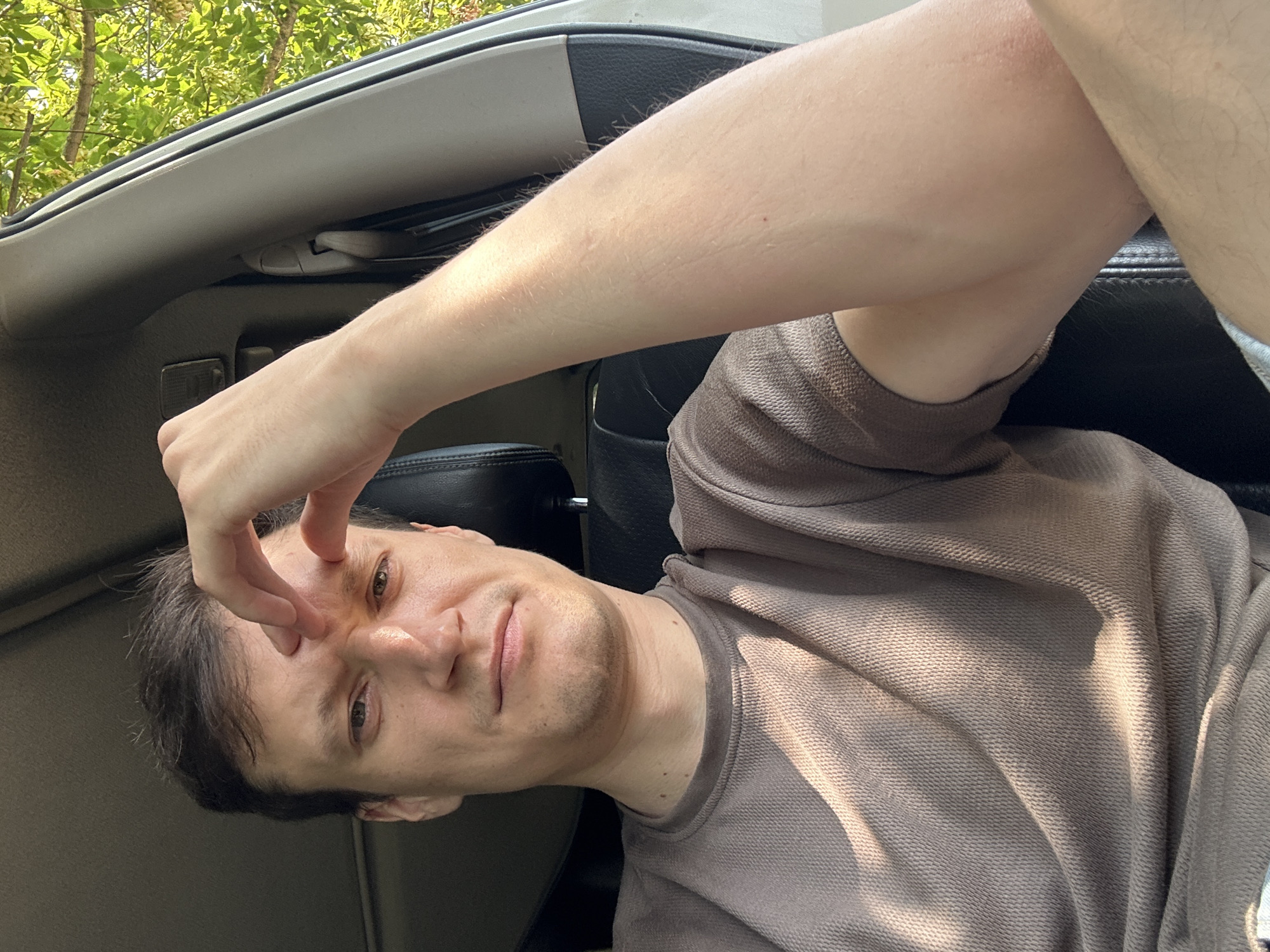 A man is seated inside a car, his right hand is touching his forehead. He is looking at the camera with a neutral expression. The man is wearing a brown t-shirt. The car interior is visible, with a black leather seat and a light-colored interior panel. Outside the car, green foliage can be seen through the window, suggesting an outdoor, possibly natural setting. The image appears to be taken in Makiv, Ukraine.