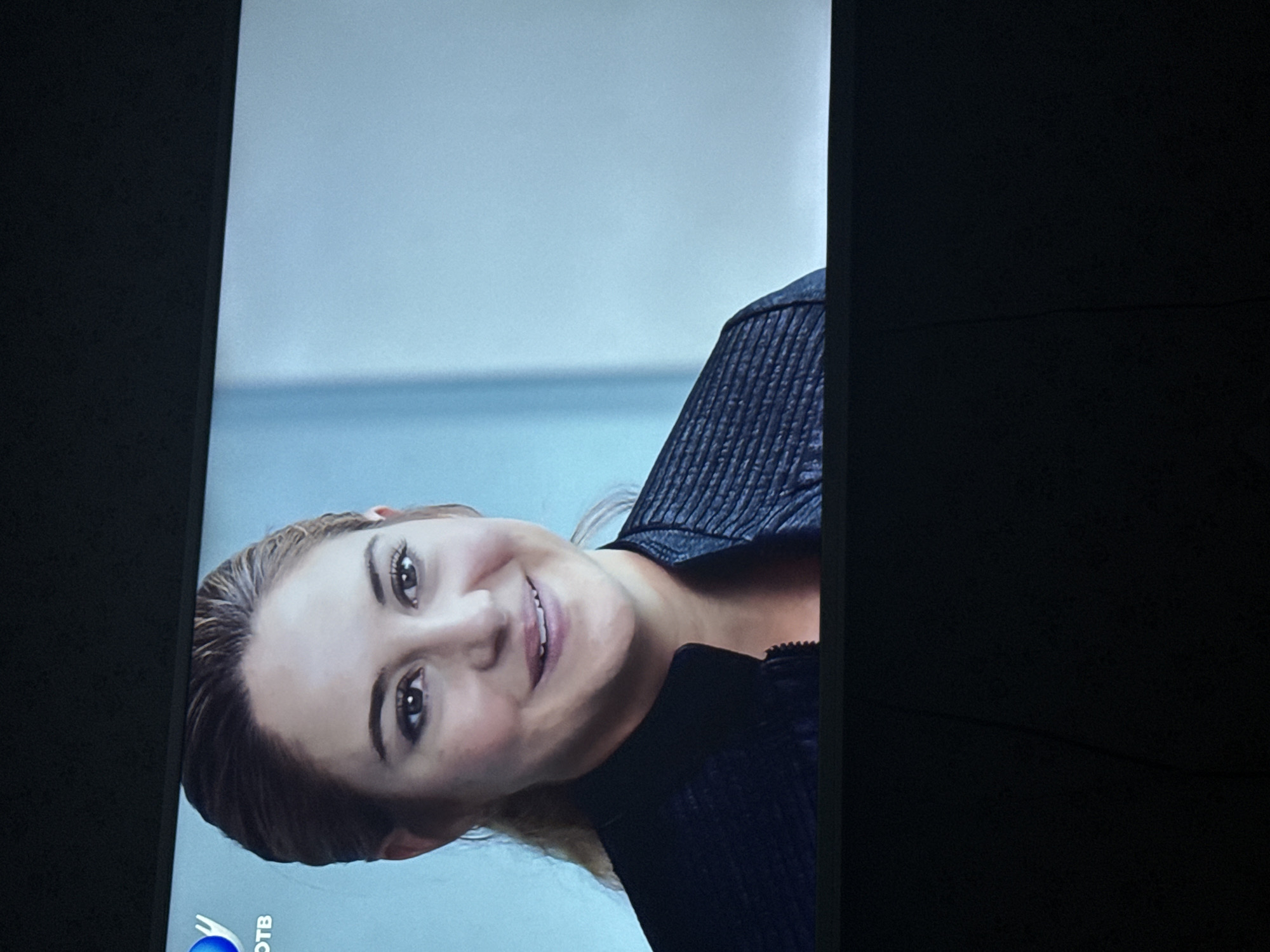 A television screen displays a woman, likely an actress, with fair skin, light brown hair, and brown eyes. She is wearing a black leather jacket. The background appears to be a light blue or gray gradient, possibly indicating an indoor setting. The lower left corner of the screen displays a logo that includes "OTB" in the lettering. The location in the city of Mytishchi, Russia is not discernible.