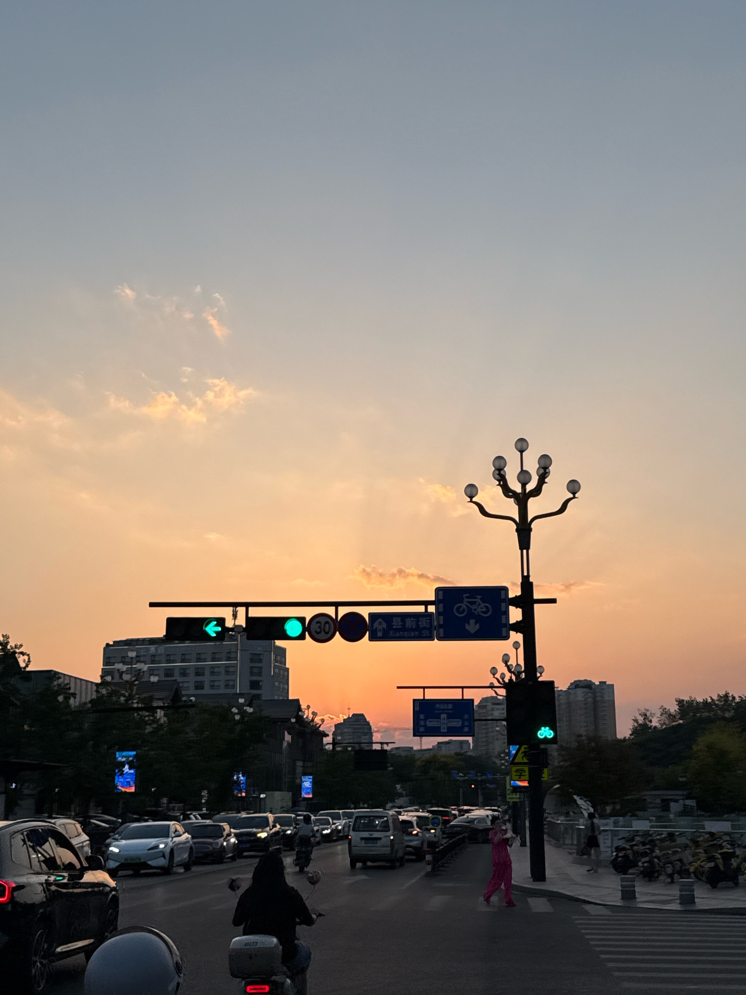 The image depicts a street scene in Yiwu, China, during sunset. The sky is a gradient of pale blue to warm orange, with scattered clouds illuminated by the setting sun. A traffic light with a green left-turn arrow and a green circular light is visible, along with a sign indicating a 30 km/h speed limit and a bicycle lane. A decorative street lamp with multiple globes stands to the right of the traffic light.
The street is congested with traffic, including cars and a few motorbikes. A person in a pink outfit is crossing the street, while another person on a motorbike is in the foreground, their back to the camera. Buildings, including an office building, line the street, and trees are visible on the sides. Several digital billboards displaying abstract images are positioned along the road. The overall impression is a busy urban environment at dusk, with the warm light of the sunset creating a specific atmosphere.