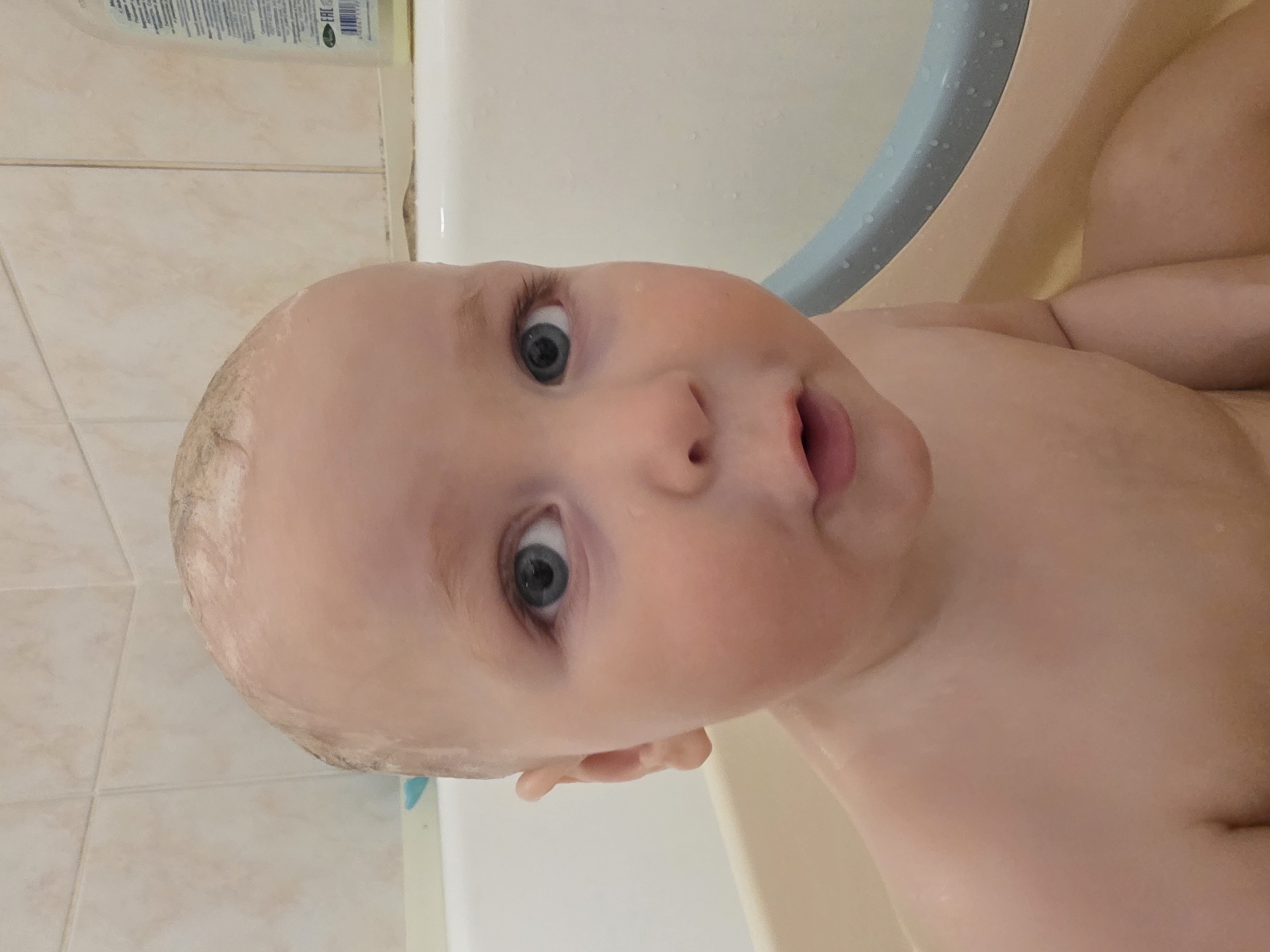 The image captures a close-up of a baby taking a bath, presented with a 90-degree clockwise rotation. The baby's face dominates the left and center of the frame, showcasing fair, soft skin and large, striking blue eyes that gaze directly towards the viewer with an expression of wide-eyed curiosity or mild surprise. The baby's mouth is slightly open, revealing a glimpse of the pink inside. The top of the baby's head shows wet, sparse light hair, and water droplets are visible on the skin, indicating immersion in water.
The upper right portion of the image shows the baby's shoulders and chest, also wet and smooth. The setting appears to be a bathtub, with its light beige or off-white surface visible around the baby. A light blue or grey plastic handle or support, possibly part of a bath seat or toy, is partially visible in the mid-right, covered in water droplets. In the background, on the left side, a light beige tiled wall with horizontal grout lines is visible. Resting on the edge of the tub or a ledge against this wall is a bottle, likely containing bath soap or shampoo, with a label displaying text. The "EAC" conformity mark is clearly legible on the label, indicating a product from the Eurasian Economic Union, likely in Cyrillic script. The lighting is bright and even, suggesting an indoor setting, possibly a bathroom, and effectively highlights the wetness on the baby's skin and the surrounding surfaces.