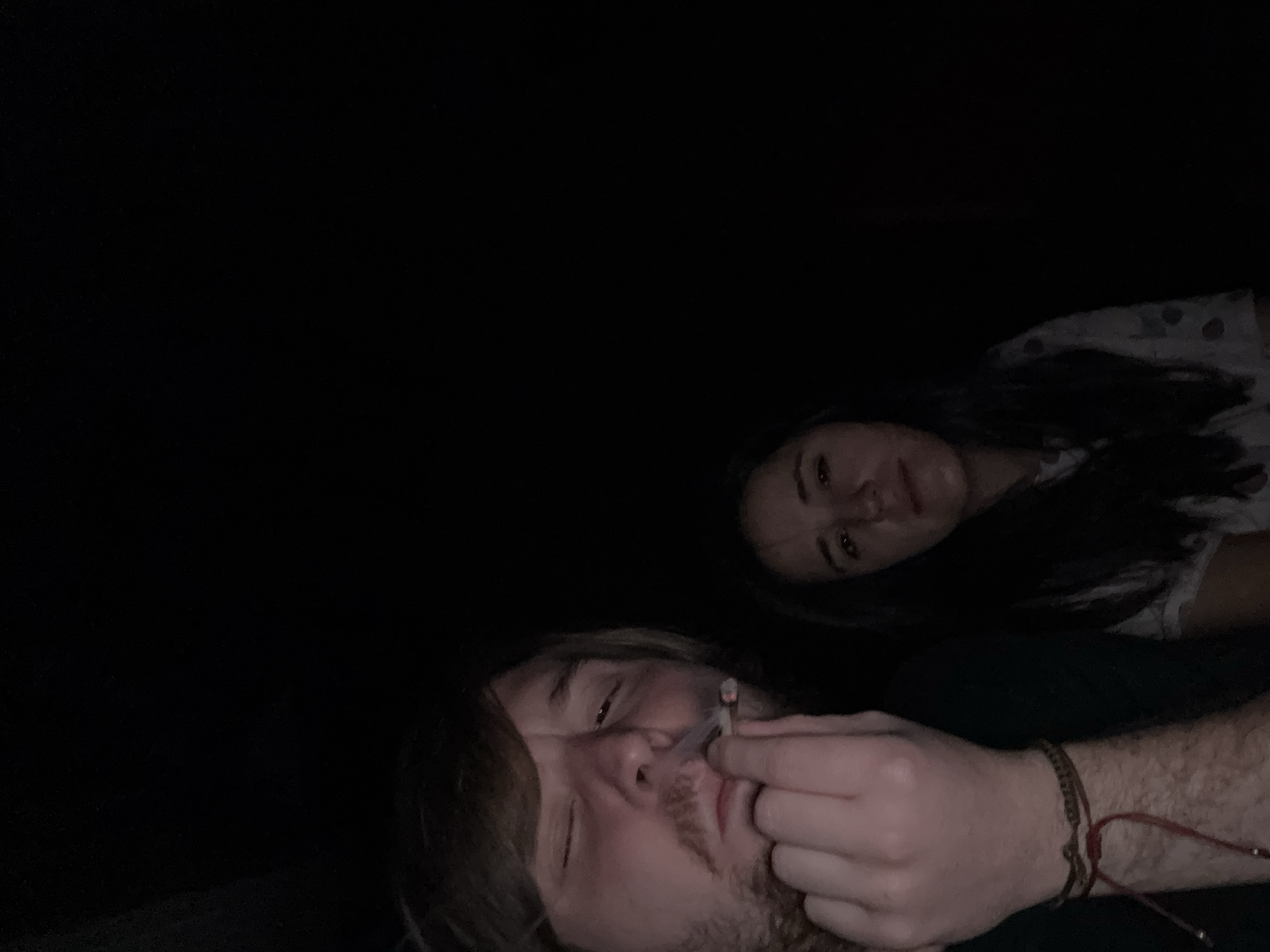 Two people are in the image. A man is in the foreground smoking. A woman is in the background. The setting is dark. The location is unknown and cannot be determined from the image.