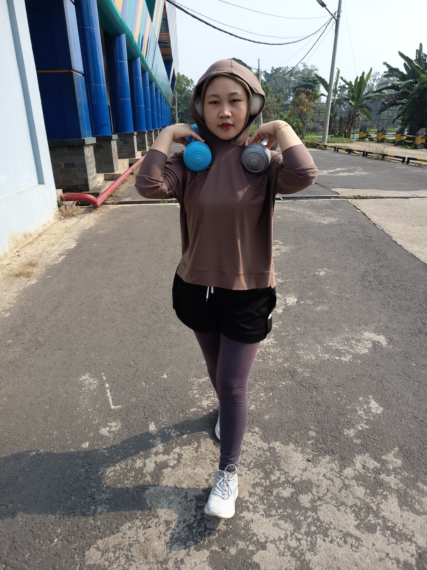 A woman with headphones around her neck, holding a pair of blue and grey dumbbells, stands on a paved path. She is wearing a brown hooded sweatshirt, black shorts over purple leggings, and white athletic shoes. The background features a building with blue and yellow striped columns, trees, and a utility pole with overhead wires. The scene appears to be outdoors, possibly in a park or recreational area in Lubuklinggau, Indonesia. The lighting suggests it is daytime.