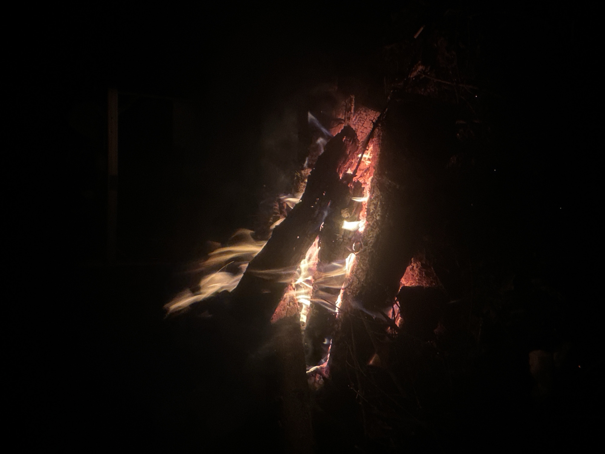 The image shows a bonfire at night. The fire is burning between several logs, with flames and smoke visible. The background is dark and unclear, but a wooden structure is visible in the left corner. The location is likely in Папярнянскі сельскі Савет, Belarus.