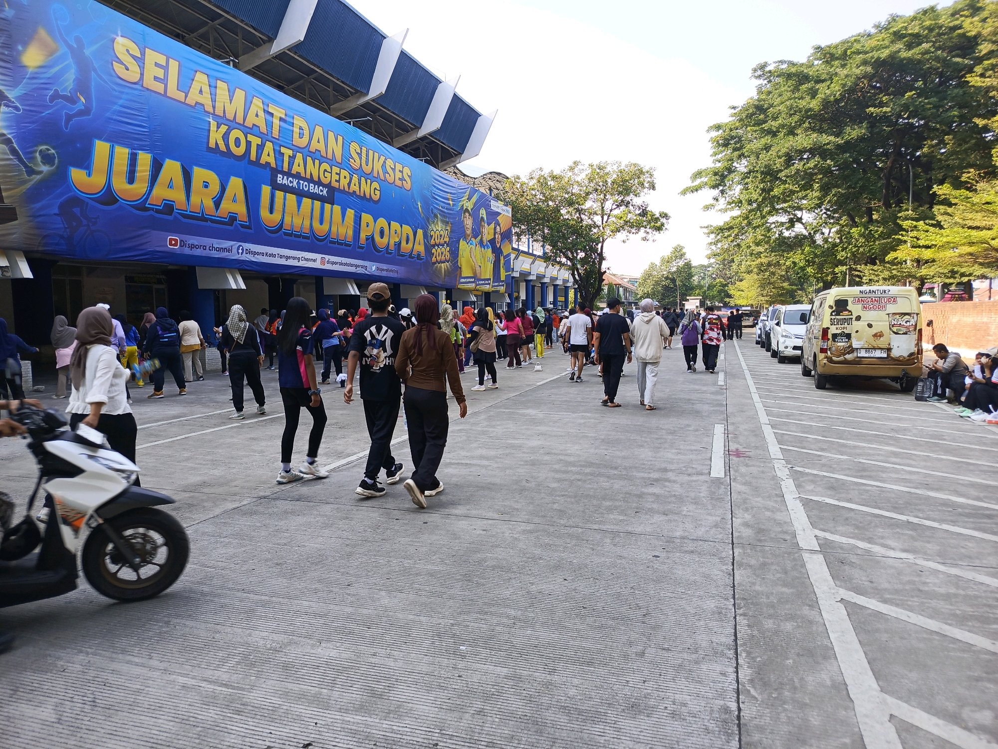 The image depicts an outdoor scene in Tangerang, Indonesia, likely at a sports stadium or public gathering area. A large blue banner with yellow text reading "SELAMAT DAN SUKSES KOTA TANGERANG JUARA UMUM POPDA BACK TO BACK" is prominently displayed on a building facade. This suggests an event celebrating the city of Tangerang's success in a sports competition. A crowd of people, dressed in casual attire, are walking and standing on a paved area. Some are wearing athletic clothing, indicating they might be participants or spectators of a sporting event. A white scooter is in motion in the foreground, partially obscuring some of the individuals. Several vehicles, including cars and a yellow van, are parked in the background. Trees and a portion of the stadium's architecture are visible. The ground is a textured concrete surface with white painted lines, possibly indicating parking spaces or a designated walking path. The overall atmosphere appears to be that of a public event or gathering.