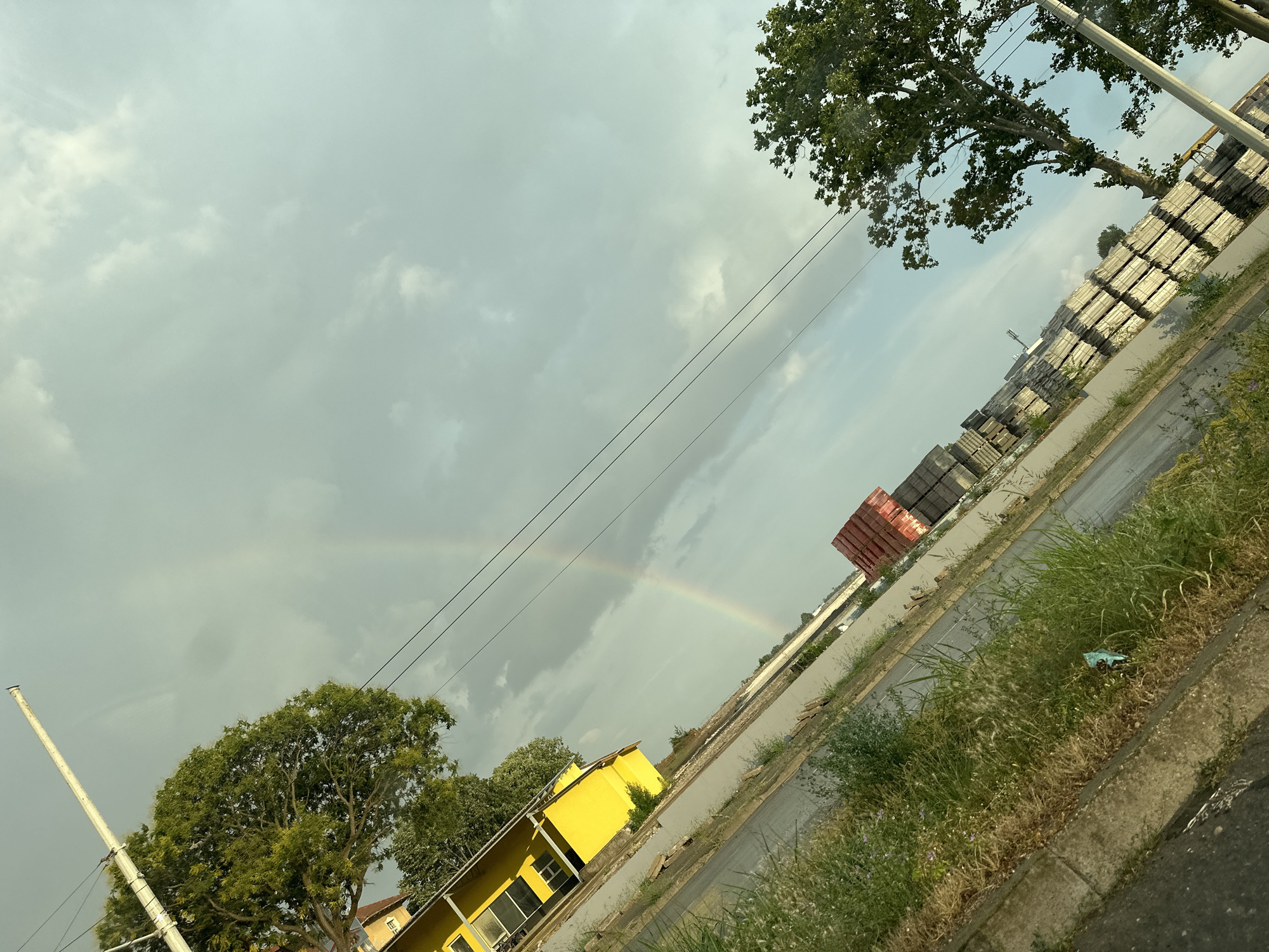 This media file depicts an outdoor scene in Ruse, Bulgaria, under a cloudy sky with a faint rainbow visible. The image is taken from a slightly tilted perspective. In the foreground, there is a grassy area with some weeds and a concrete curb. A road runs diagonally through the frame, with a yellow building on the left side. Beyond the road, there is a large pile of grey blocks, possibly for construction, and further in the distance, more stacks of what appear to be building materials or containers. Two sets of power lines run across the sky. A large tree with green foliage is prominent in the upper right corner, and another smaller tree is visible near the yellow building. The time of day appears to be late afternoon or early evening, given the lighting conditions and the clouds. The presence of the rainbow suggests recent or ongoing rain. The scene conveys a sense of everyday life with elements of infrastructure and nature. No people or text are clearly visible in the image.