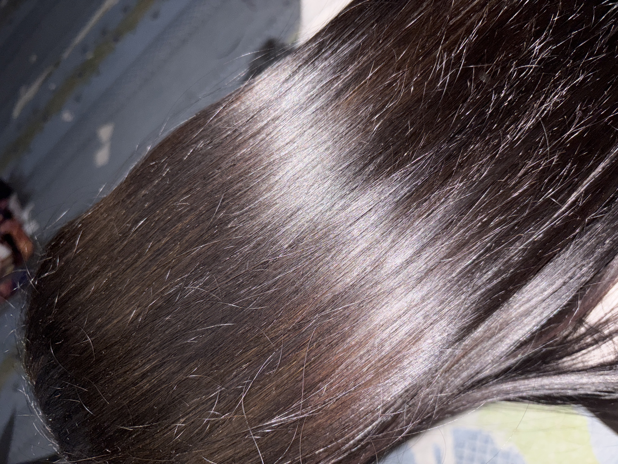 A close-up shot displays a section of dark brown, shiny hair. Individual strands are visible, with variations in tone ranging from lighter brown to almost black. The hair appears smooth and well-maintained, reflecting light in a way that accentuates its texture. The hair rests on a surface. The location cannot be ascertained based on the image.
