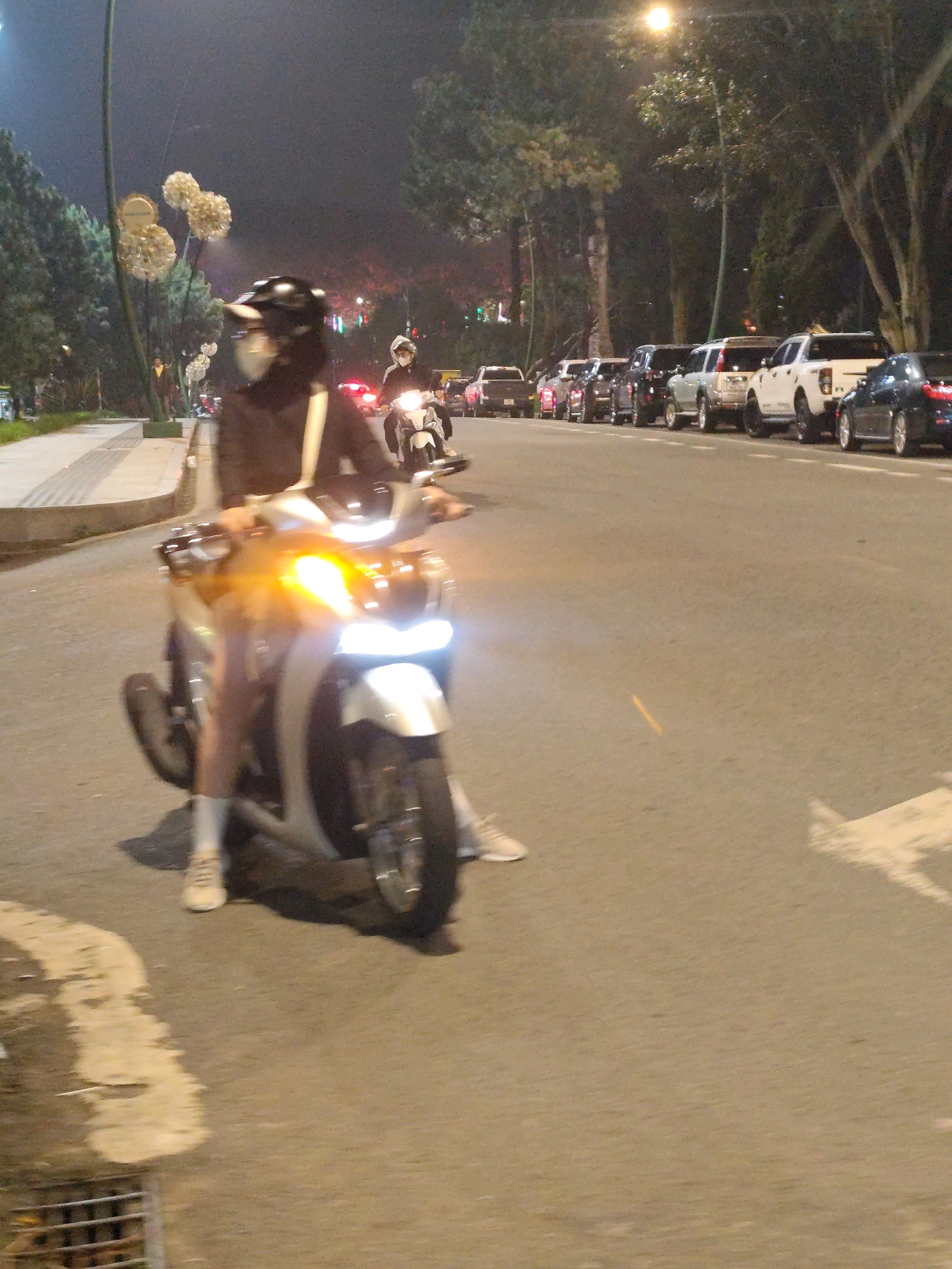 The image depicts a street scene at night in Da Lat, Vietnam, featuring two individuals on motorcycles. In the foreground, a person on a white scooter is visible, with their legs and feet clearly showing, wearing white socks and light-colored shoes. The scooter's headlight is illuminated, casting a bright orange glow. The rider is wearing a dark jacket and a helmet with a mask. In the background, a second person on a motorcycle, also wearing a helmet, is seen further down the road. The street is lined with parked cars, including several SUVs and a dark sedan. Trees are present along the side of the road, and a sidewalk with decorative streetlights is visible on the left. The overall lighting suggests it is nighttime, with artificial lights from streetlamps and vehicles illuminating the scene.