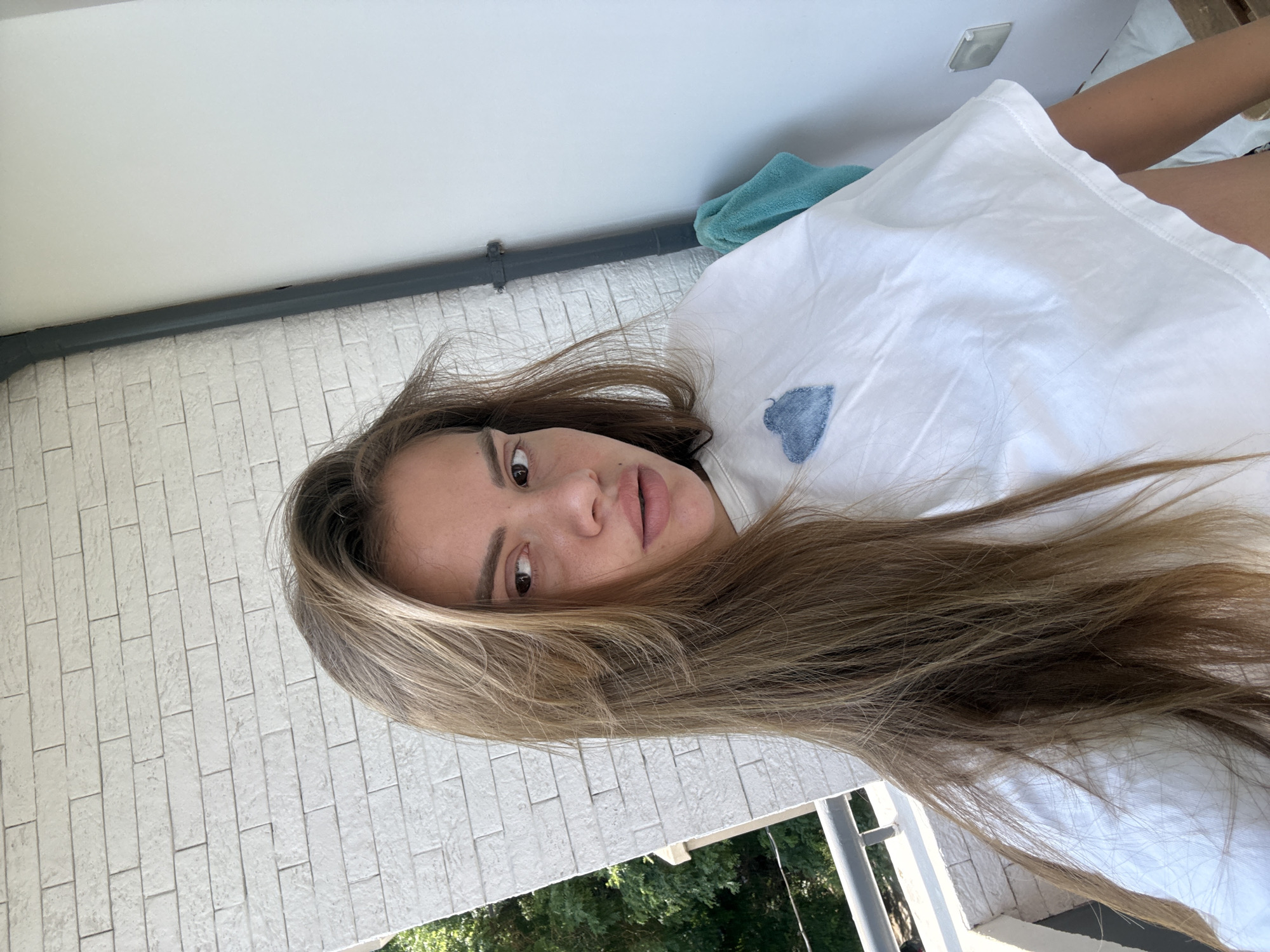 The image, rotated 90 degrees counter-clockwise, captures a young woman lying down and looking directly at the camera. She has long, wavy brown hair with lighter highlights, brown eyes, and full, slightly parted lips, conveying a relaxed expression. She is wearing a white top or dress, and part of her bare leg is visible towards the right side of the frame.
She rests on a white surface, possibly a pillow or blanket, which features a small, light blue, heart-shaped patch made of denim-like fabric. Behind her head, a folded teal or turquoise towel is visible.
The setting appears to be a brightly lit room or balcony, characteristic of Plovdiv, Bulgaria. Above her head (to the left in the rotated image), a plain white ceiling with a small light fixture is visible. A dark grey pipe or rail runs horizontally along a wall line. Below this, the wall is clad with white, rectangular, tile-like bricks. Through what seems to be a window or an open space below the tiled wall, vibrant green foliage from trees and a white structural element, possibly a balcony railing, are seen, suggesting an outdoor view. The lighting is bright and natural, indicating daytime. There is no visible text in the image.