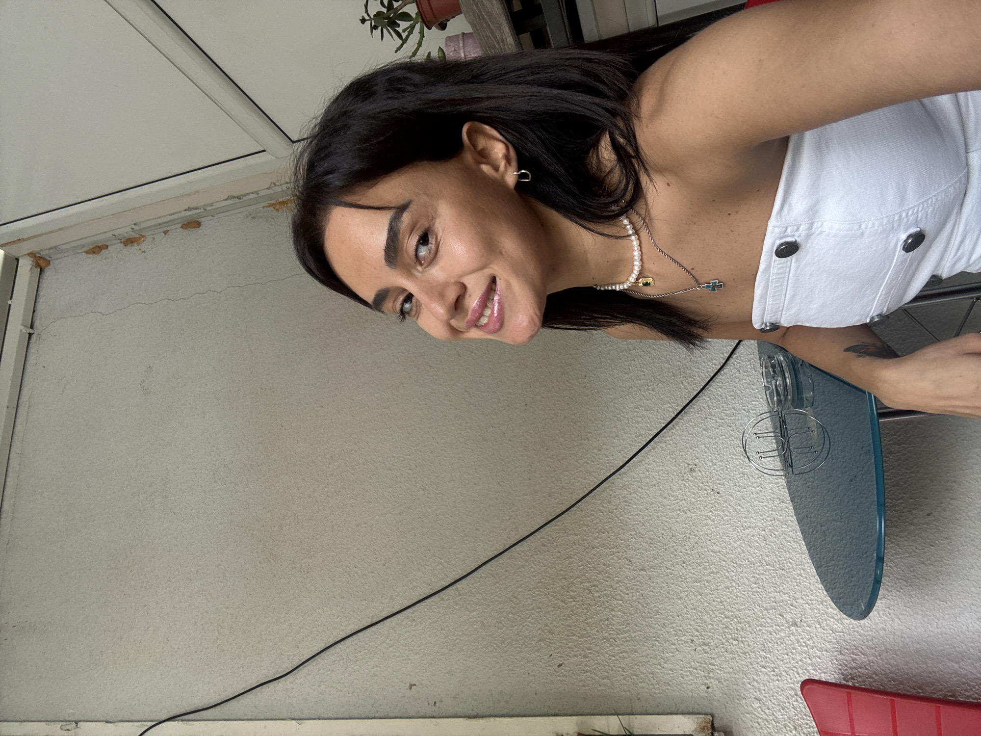 The image is a selfie taken outdoors, possibly on a balcony or patio, with a textured, light-colored wall behind the subject. The subject is a woman with dark hair and dark eyes, wearing a white strapless top and a pearl necklace with a pendant. She is smiling and looking towards the camera. A small round glass table with an ashtray and a metal object on it is visible in the foreground. A red chair is partially visible on the left side of the image. In the background, there is a potted plant on a shelf. The overall scene suggests a casual setting, possibly a cafe or a residential outdoor space in Tbilisi, Georgia.