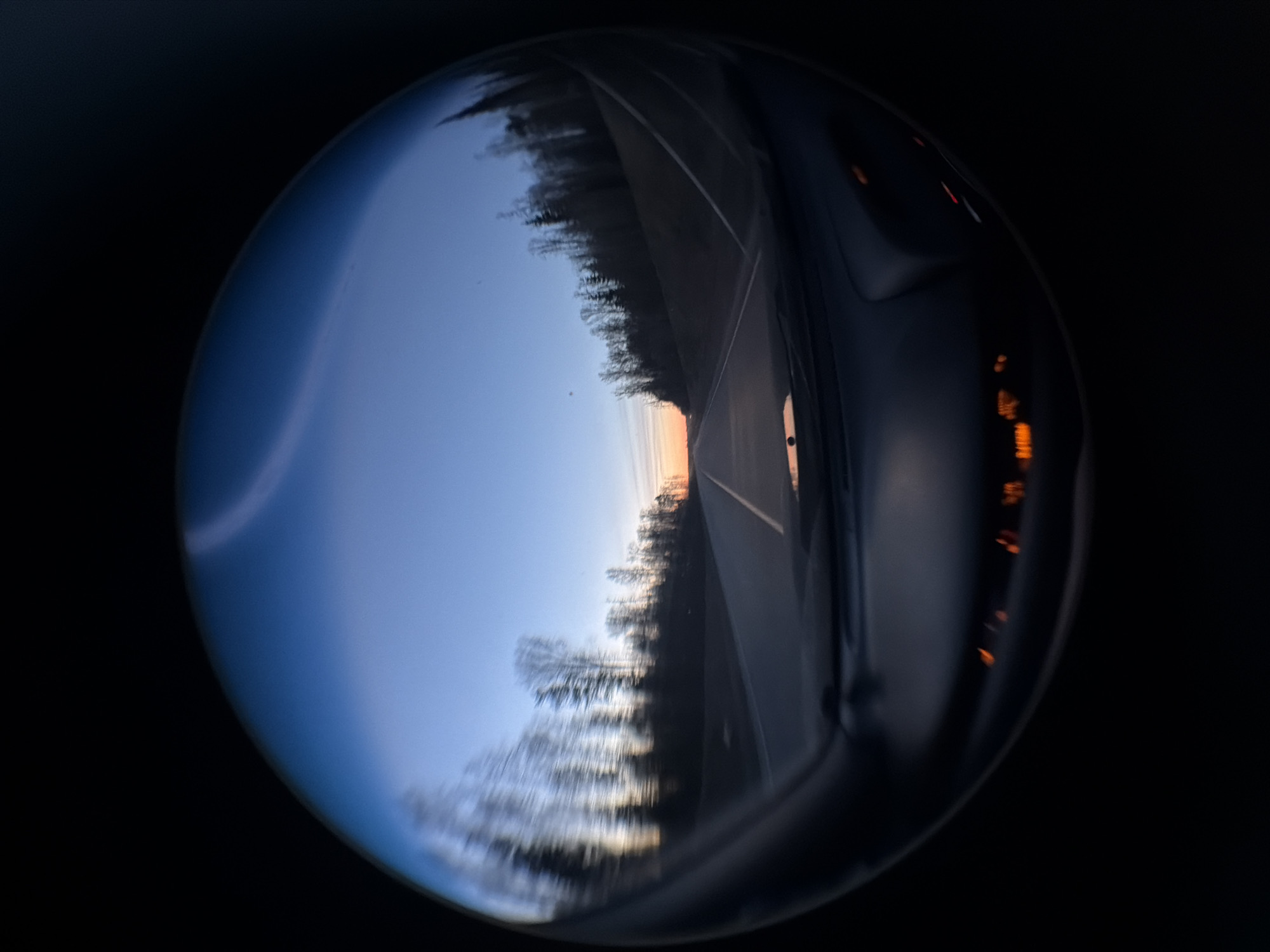 The image appears to be a reflection in a convex mirror or lens, presenting a distorted view of a highway at twilight. The highway has a central yellow line. Trees line the side of the road. There are reflections of an interior car dashboard. The environment is generally dark, suggesting dim lighting or night.