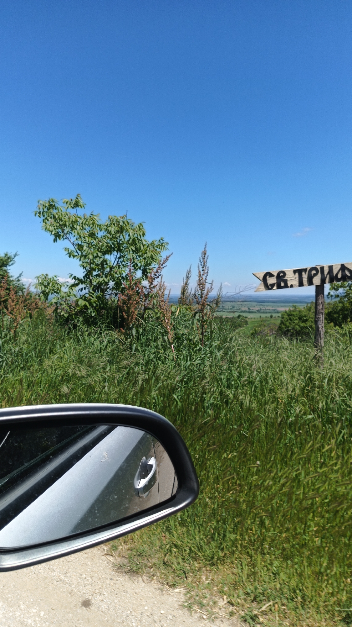 The image depicts a view from a car, likely in a rural or semi-rural setting. A car's side mirror is visible in the foreground, showing a reflection within. Outside the car, there's lush, green vegetation including tall grass and some bushes, obscuring the view to some extent. A wooden sign with lettering, potentially indicating a location or direction, is partially visible amidst the vegetation. Beyond the greenery, the landscape opens up to a valley or plain, with a clear blue sky overhead.
The sign seems to say "СВ. ТРИАДА" in Cyrillic alphabet. This would likely be "Sveta Triada" which means Holy Trinity, a common name for a church or place in Bulgaria.
Given the location of Gornoslav, Bulgaria, it is presumed that the image was captured in the immediate vicinity. No people, interactions, or actions are explicitly visible in the frame.