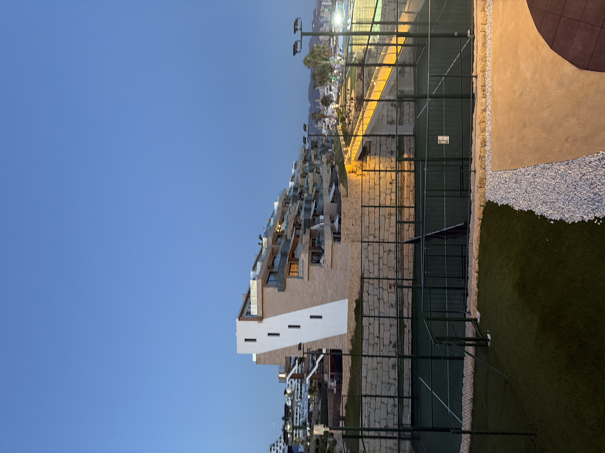 The image depicts an outdoor scene in Finestrat, Spain, at dusk. The foreground features a padel tennis court with green turf and white markings, enclosed by a green metal fence. Adjacent to the court is a patch of artificial grass and a pathway made of white gravel leading to a concrete area. In the mid-ground, a stone retaining wall supports a tiered residential building with a distinctive white slanted facade and multiple balconies. Beyond the building, more residential structures are visible on a hillside, along with palm trees and distant mountains. The sky is a clear, deep blue, transitioning to lighter shades near the horizon. Lighting fixtures on poles illuminate the scene. No people are visible in the image.