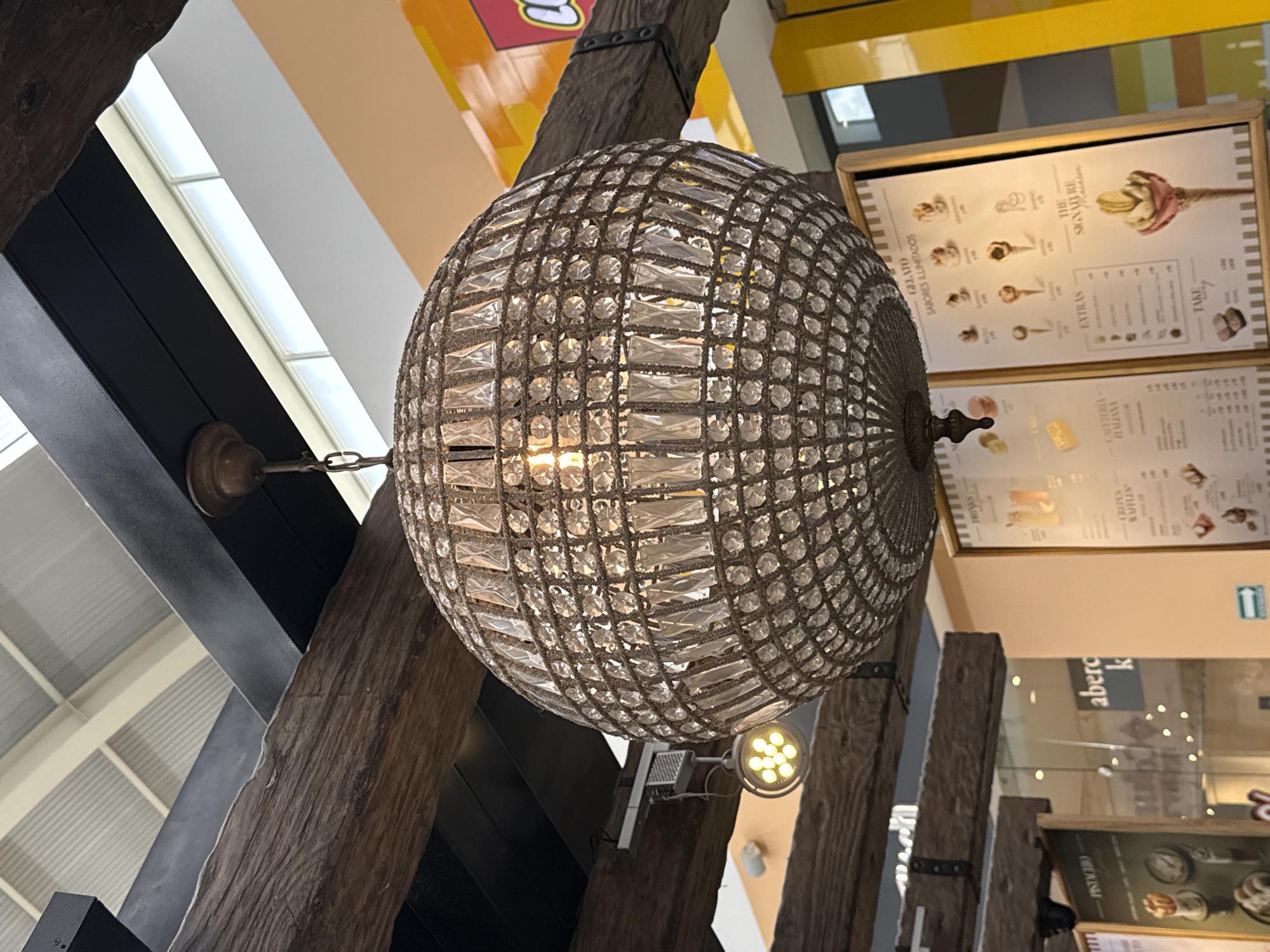 The image shows an ornate, globe-shaped chandelier with a crystal-like appearance hanging from a wooden beam structure. The chandelier appears to be in an indoor setting, possibly a commercial or retail space. Behind and to the right of the chandelier are menus displaying gelato flavors, mounted on a wall. Further in the background, indistinct signage is visible above a glass partition that reads "aberc". The environment seems to be a shopping center or mall. The photo is taken from a low-angle perspective. The location is not specifically determinable as San Francisco Ocotlán (Ocotlán), Mexico based on visual information alone.