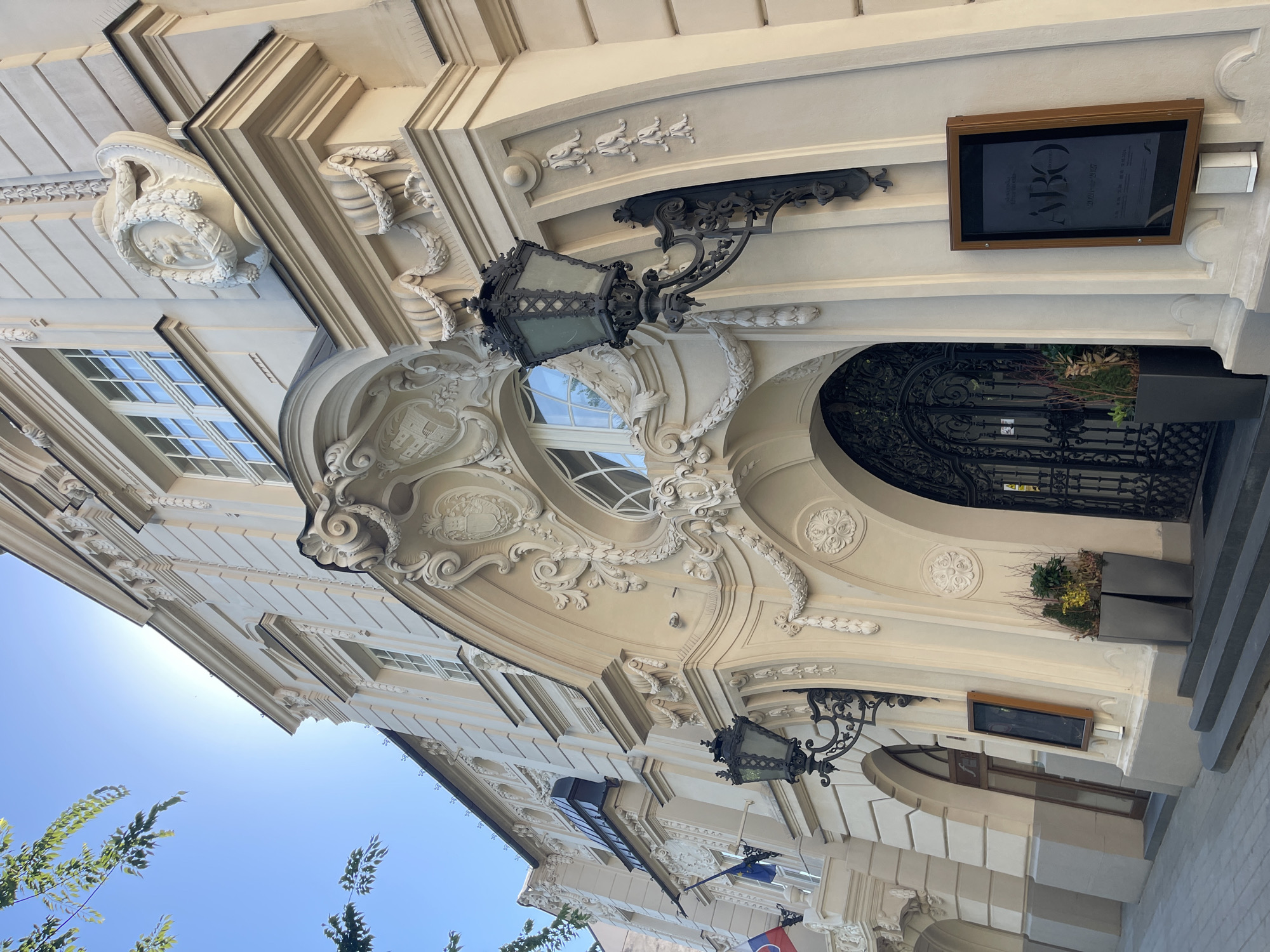 The image shows the facade of a building in Bratislava, Slovakia, likely a historical building due to its ornate architectural style. The building is light-colored, possibly sandstone or stucco, and features intricate decorative elements such as carvings, garlands, and urns. The main entrance is a large, arched doorway with decorative black metalwork. Flanking the entrance are large, wrought-iron lamps. Two flags are visible on the left side of the building, one of which appears to be the Slovak flag. A sign is mounted on the wall to the right of the entrance, displaying text and graphics, possibly indicating an exhibition or event. There are planters with greenery near the entrance. The sky is bright blue, suggesting a clear day, with some branches of a tree visible in the upper left corner. The perspective is from a low angle, looking up at the building.
