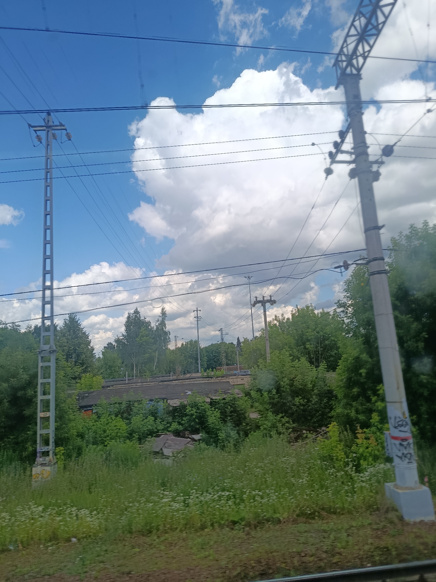 The image shows a view from a moving vehicle, likely a train, looking towards a landscape with power lines and vegetation. In the foreground, there are power lines stretching across the frame. Two tall, grey metal utility poles are visible, one on the left and one on the right. The pole on the left has a rectangular lattice structure, while the pole on the right is a single, thicker concrete pole with some graffiti on it.
In the middle ground, there are dense trees and bushes, with some buildings or structures partially visible behind them. The buildings appear to have dark, flat roofs and are somewhat obscured by the foliage. The vegetation is lush and green, indicating a warm season.
The background consists of a bright blue sky with fluffy white clouds. The overall impression is of a rural or semi-rural area, possibly on the outskirts of a city, with infrastructure related to power transmission. The location is identified as Минвуз Снт, Russia. There are no people visible in the image.