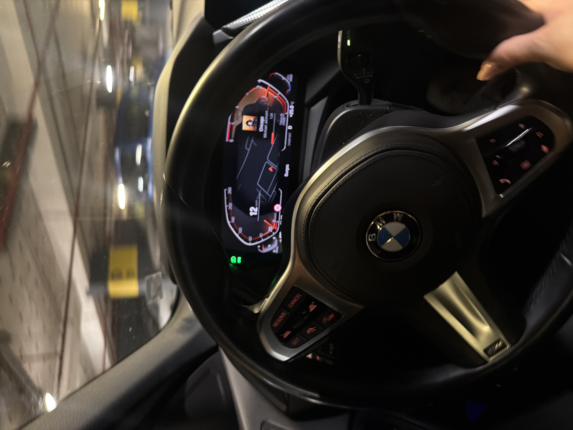 An interior shot of a car's dashboard and steering wheel shows a digital display indicating "Chicago," "Michael Jackson," and "1:24." The car is likely in a parking garage, suggested by the concrete ceiling and lighting visible through the windshield. A hand with painted nails is visible on the right side of the frame, near the steering wheel. The car is a BMW, indicated by the emblem on the steering wheel. The image appears to be taken while stationary, as evidenced by the parked car interior and the stable focus on the dashboard.