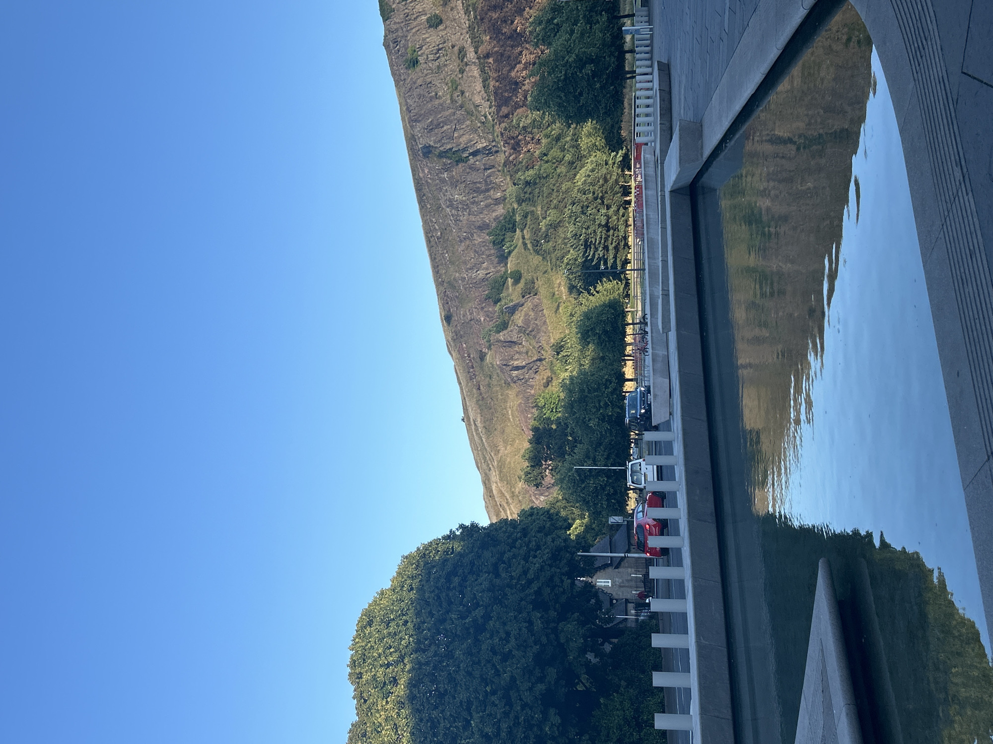 This image captures a serene daytime scene in the city of Edinburgh, United Kingdom, likely near Holyrood Park. In the foreground, a modern water feature with clean, geometric lines reflects the clear blue sky and the verdant slopes of a prominent hill in the background. The hill's rocky facade and scattered vegetation are visible, illuminated by natural light. Beyond the water feature, a paved area with concrete benches and bollards leads to a road. Cars, including a red hatchback, a white van, and a dark SUV, are parked or driving on the road. A large, mature tree with dense foliage dominates the left side of the frame, partially obscuring a small building with white-trimmed windows. The overall atmosphere is tranquil, with the bright sky and the stillness of the water suggesting a peaceful day in the city.