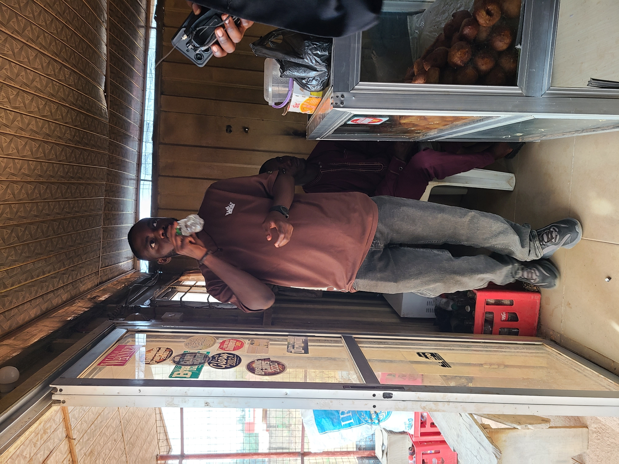 A man in a brown shirt and gray pants is standing in what appears to be a small food stall in Gura Top, Nigeria. He is drinking from a plastic bottle. Behind him, another man is seated on a white plastic chair. The stall has a glass display case with stickers on it, and a metal food warmer containing donuts sits on top. A person is holding a cell phone near the food warmer, possibly taking a picture. Crates of soda bottles can be seen near the stall's base. The stall is enclosed with corrugated metal panels and has a window offering a view of the outside.