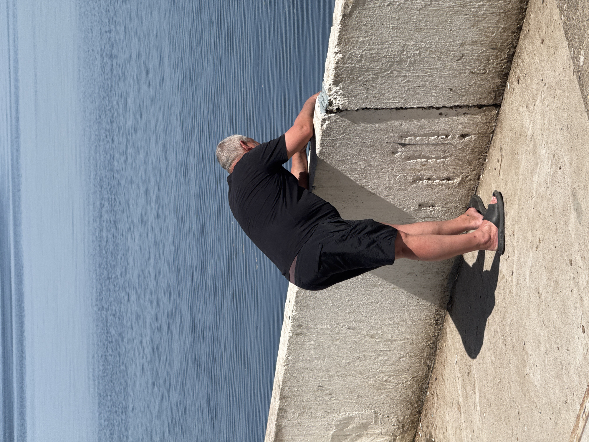A man with short gray hair wearing a black T-shirt and black shorts is standing on a concrete ledge next to a body of water. He is holding onto the ledge with his hands. The location is Unknown, Unknown.