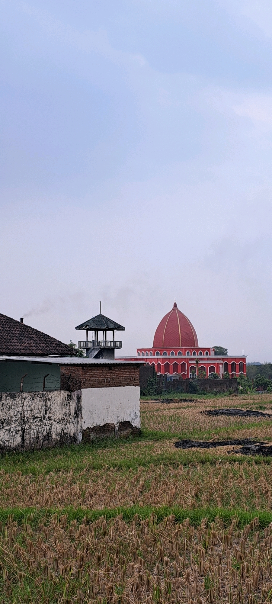 The image depicts an exterior view of the Ketanireng Grand Mosque in Ketanireng, Indonesia. The mosque is characterized by its prominent red dome and is situated in a rural or semi-rural landscape. In the foreground, a low, weathered wall constructed of white plaster and brick is visible, partially obscuring a tiled roof structure on the left. To the right of this wall, a tall, slender observation tower with a green, multi-sided roof stands. Beyond the observation tower and the wall, the mosque's distinctive red dome and arched facade dominate the middle ground. The surrounding area appears to be a field of dry grass or stubble, suggesting it might be a harvested agricultural area. The sky is overcast and pale. There are no people or animals visible in the scene.