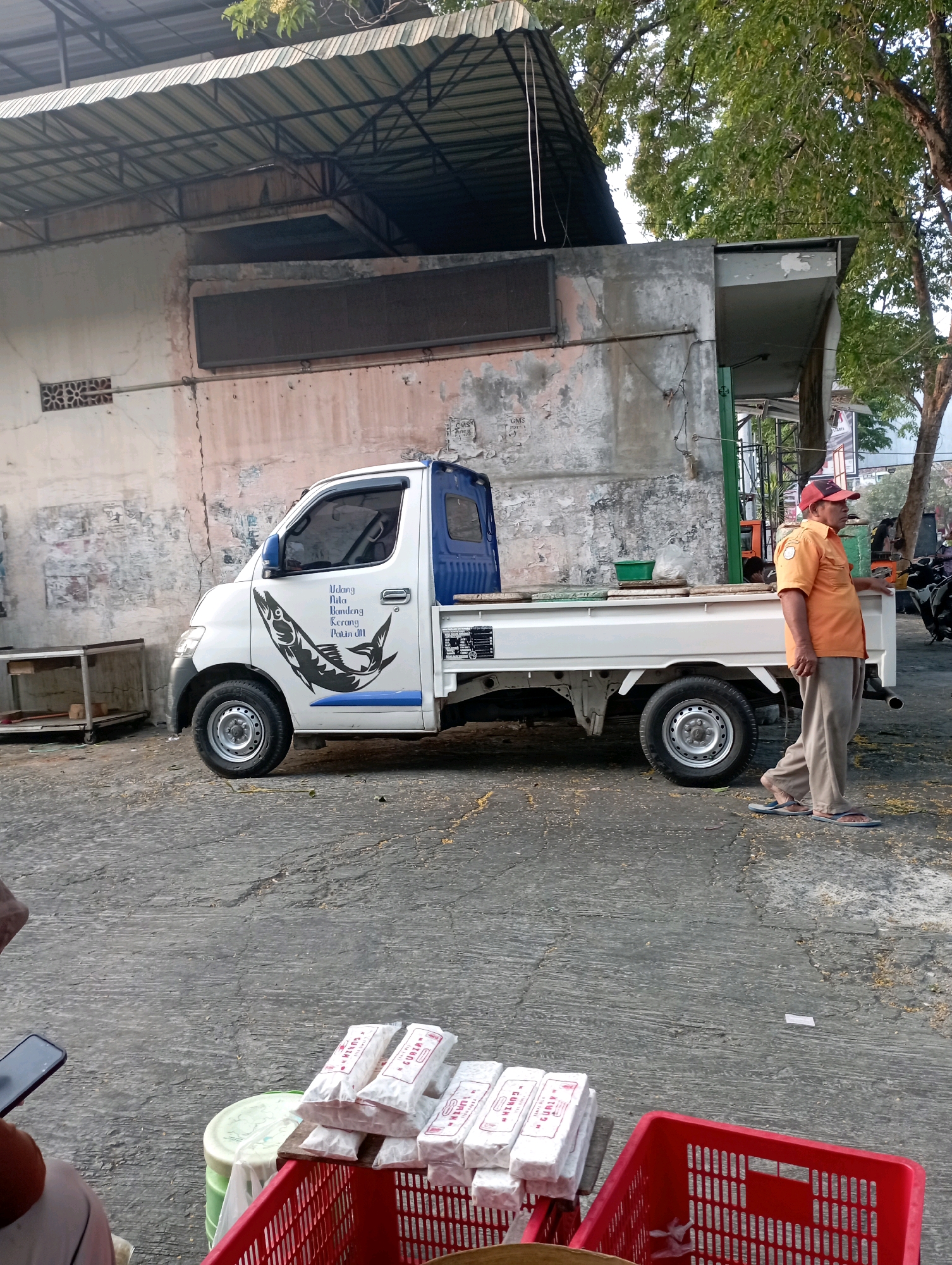 A white pickup truck with a blue cab is parked on a paved surface in front of a weathered building with a corrugated metal awning. The truck has a decal of a fish on its side and text in blue lettering. A man wearing an orange shirt, khaki pants, a red cap, and flip-flops stands to the right of the truck, looking to his left. In the foreground, there are several white packages of what appear to be food items stacked on a wooden board and a red plastic crate. The environment appears to be an outdoor market or roadside stall. The scene is likely located in Ponorogo, Indonesia, given the text on the truck and the presence of items commonly found in Indonesian markets.