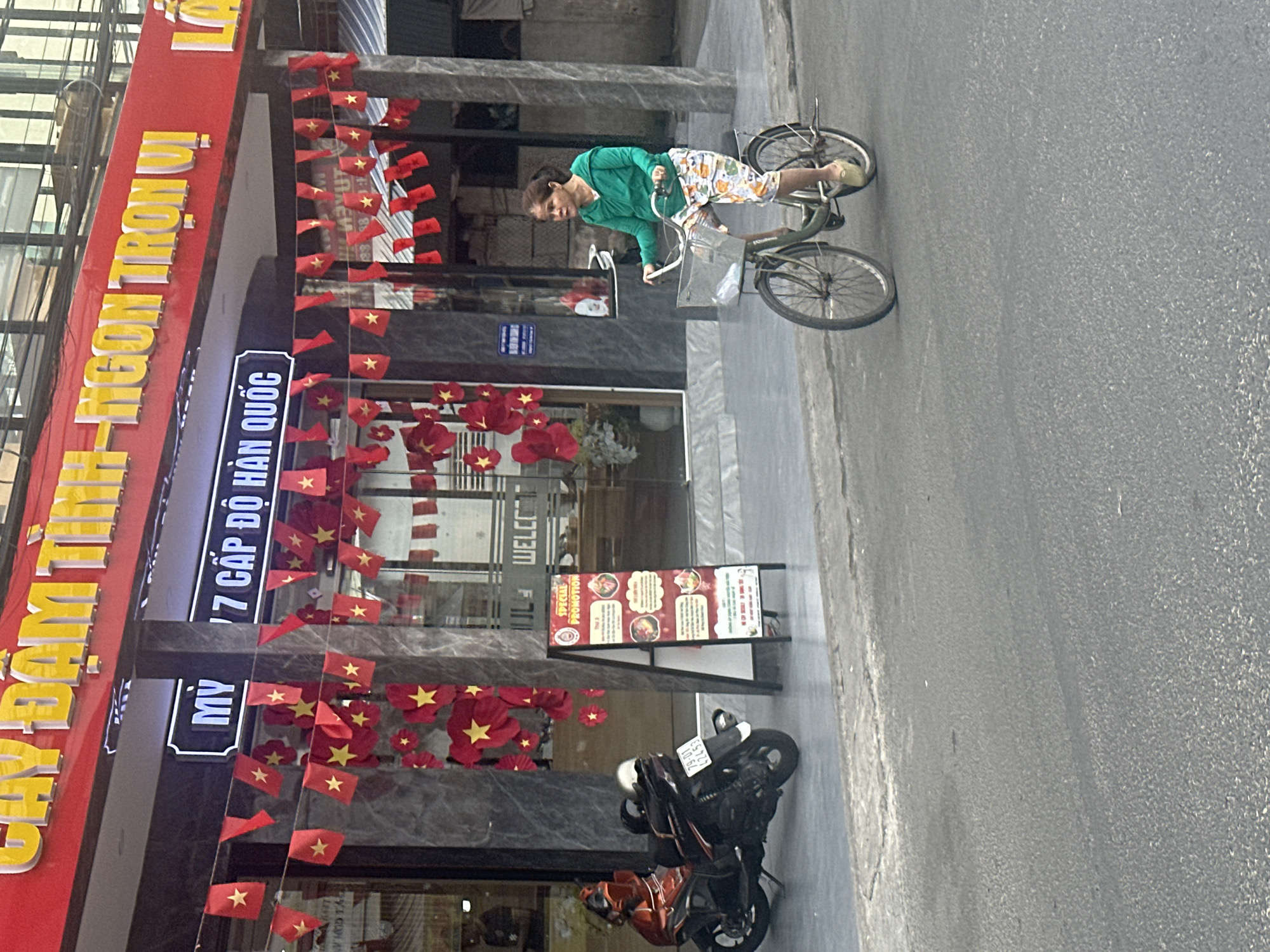 The image captures a street scene in Bac Nha Trang Ward, Vietnam. A woman in a green shirt and patterned shorts is riding a bicycle on the right side of the frame, moving away from the camera. To the left, a restaurant or business establishment is adorned with Vietnamese flags. A sign in front of the building reads "MỸ 17 CẤP ĐỘ HÀN QUỐC," and a smaller sign advertises a "SPECIAL PROMOTION." A parked motorcycle is visible near the building. The environment appears urban.