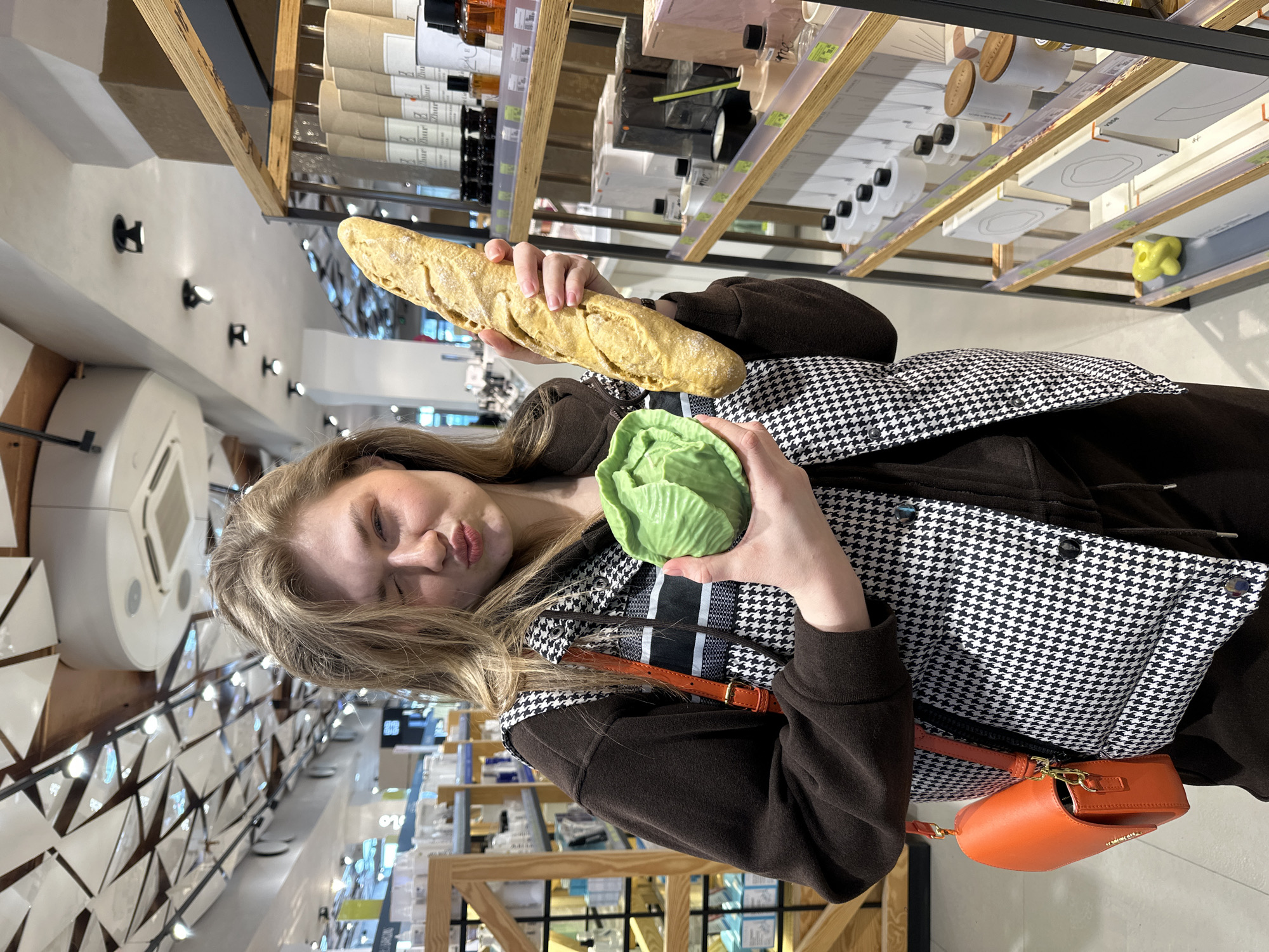 An image captures a young adult female inside a Zhur retail store, identified by product packaging, located in Kazan, Russia. The woman, with light hair, is positioned centrally, facing the camera with a wink of her right eye and puckered lips. She wears a dark brown long-sleeved top beneath a black and white houndstooth puffer vest, with an orange shoulder bag visible on her right hip. In her left hand, she holds a light green, cabbage-shaped novelty item, and in her right hand, she holds a golden-brown, baguette-shaped novelty item; both resemble bath or soap products. The background reveals rows of wooden shelving displaying various merchandise, including cylindrical "Zhur" branded packages, glass bottles, diffusers, and boxed goods. The store's ceiling features a distinctive modern design with geometric white panels and integrated spotlights.