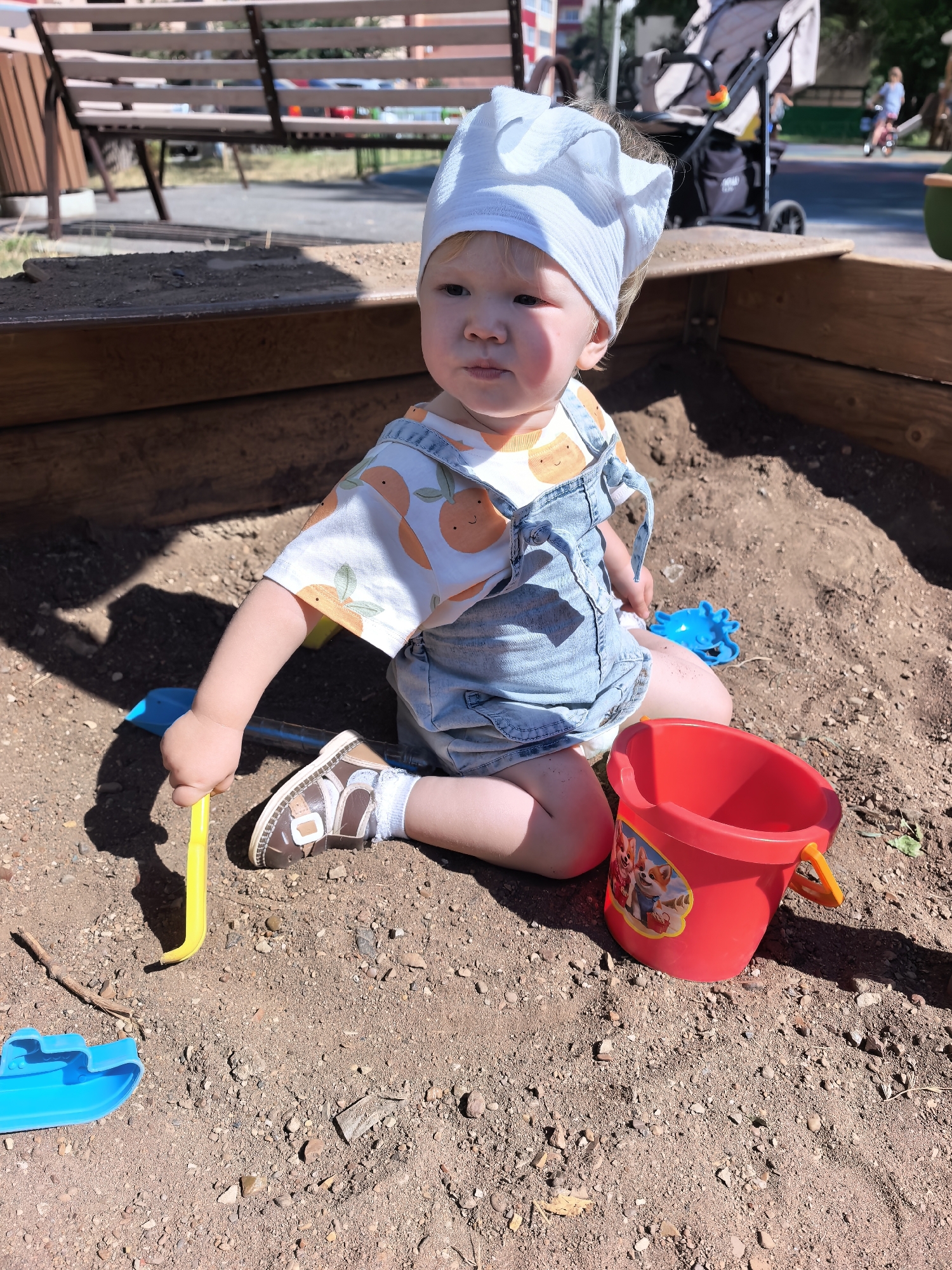 A young child is playing in a sandbox. The child is wearing a white head covering, a white shirt with an orange fruit pattern, and blue denim overalls. The child is holding a yellow toy shovel. Next to the child is a red bucket with a Paw Patrol design and a blue toy boat. The sandbox is made of wood and filled with sand. In the background, there is a wooden bench and a stroller. This scene is taking place in a public park in Sterlitamak, Russia, evidenced by the presence of children's play equipment and typical park elements.
