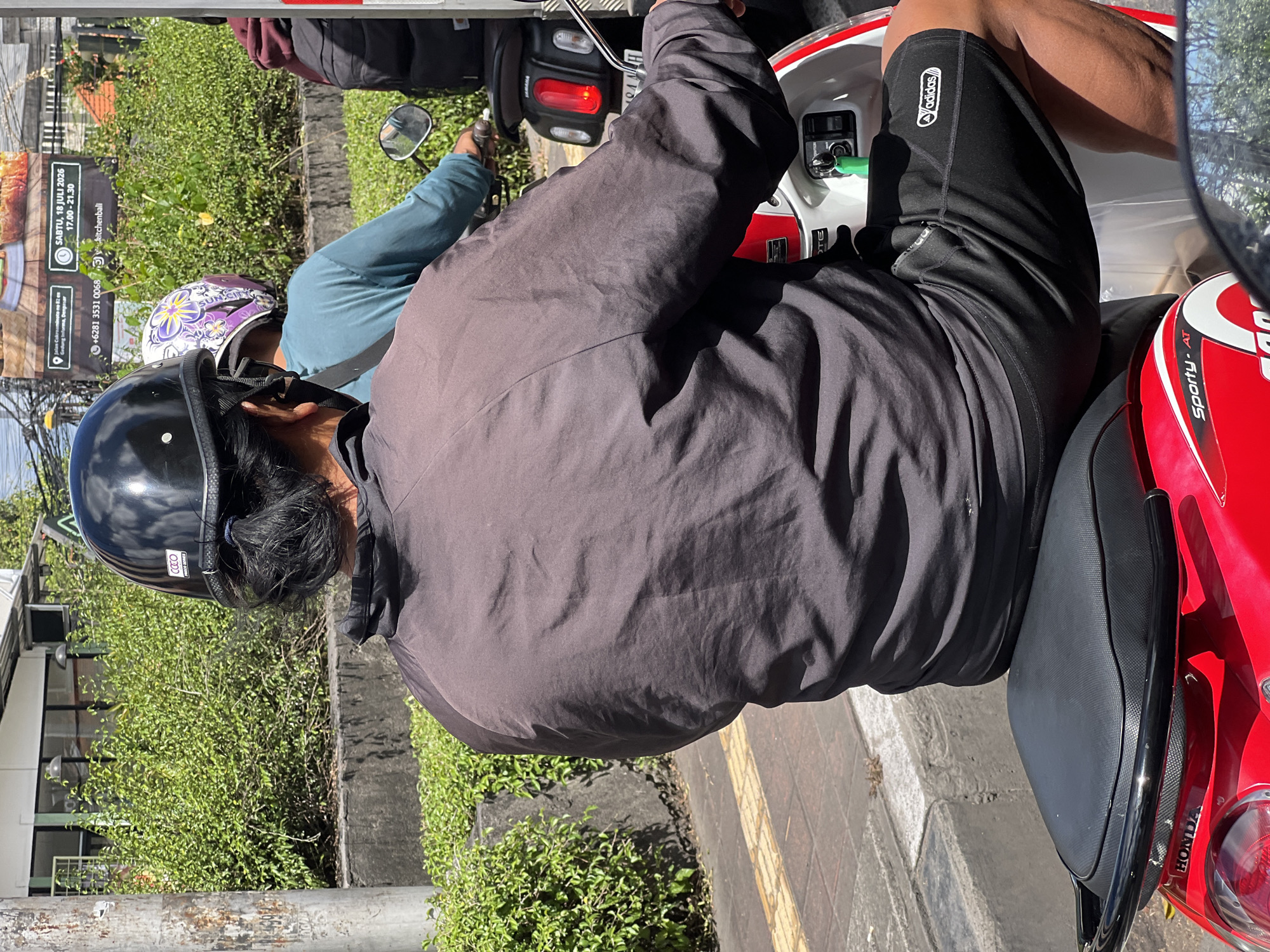 This image captures a street scene in Denpasar, Indonesia, with a focus on individuals riding motorcycles. In the foreground, a person wearing a black helmet and a dark jacket is riding a red and white motorcycle. The person's hair is tied back in a ponytail. The jacket appears to be made of a light, possibly water-resistant material. The person is also wearing black shorts with the Adidas logo visible on the right leg.
Behind the person in the foreground, another individual wearing a blue shirt and a helmet with a purple floral pattern is riding a motorcycle. Further back, a third person, partially visible, is wearing a dark hooded jacket and a backpack. Both the second and third individuals appear to be on the same motorcycle, suggesting a tandem ride.
The environment consists of a street with a sidewalk and lush green bushes lining the side. A concrete barrier separates the road from the sidewalk. A pole is visible on the left side of the image, and a sign with text in Indonesian is displayed in the background, indicating the location as Denpasar. The sign includes information about a kitchen and a date, "Sabtu, 18 Juli 2026". The lighting suggests it is daytime, with sunlight casting shadows. The overall scene depicts typical urban traffic and daily life in Denpasar.
