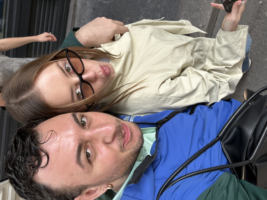 A high-angle, close-up photograph captures two adults, a male and a female, posing in an outdoor setting in Paris, France. The male, on the left, has dark, curly hair, light-colored eyes, and minimal facial hair, including a subtle mustache and goatee. He wears a light green collared shirt beneath an open royal blue and dark green color-blocked jacket. His left ear features a gold hoop earring and a small stud. A black, structured tote bag hangs across his lower torso. The female, positioned on the right, has straight, light brown hair parted down the middle. She wears black cat-eye sunglasses with amber-tinted lenses and a light beige, long-sleeved oversized jacket. Her lips are pursed, and her right arm is draped over the male's left shoulder. She wears rings on her right hand, which holds a dark-colored smartphone or similar device. Both individuals are looking directly at the camera. In the blurred background, the lower torso and right arm of a third person, wearing a light grey garment and a ring, are partially visible against an indistinct grey surface, likely pavement or a wall.