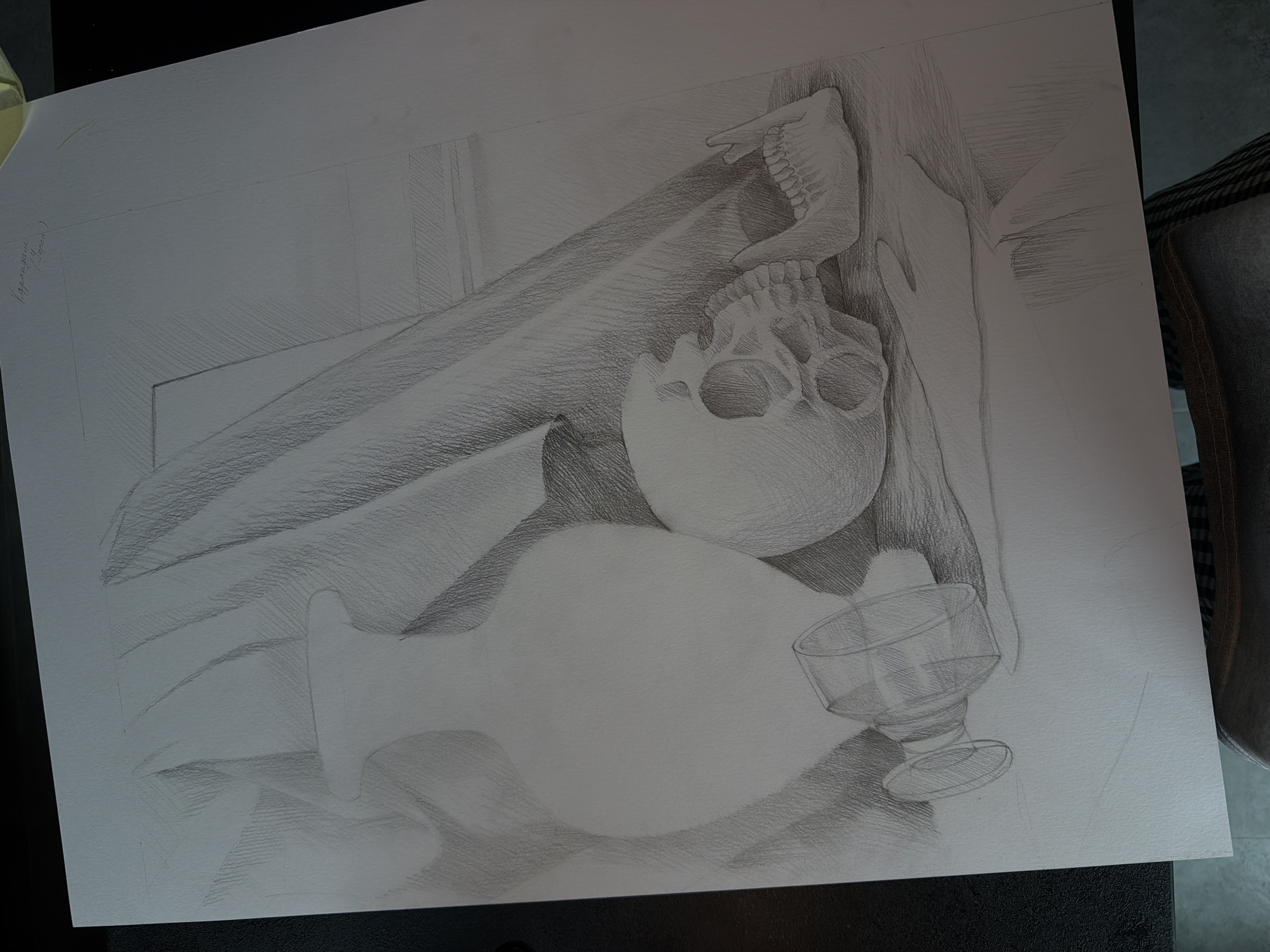 The image is a pencil drawing of a still life. The subject matter includes a human skull with a detached jawbone resting on top, a large vase with a bulbous shape, and a stemmed glass. These objects are arranged on what appears to be draped fabric. The background includes angular structures, possibly wooden panels or planks. The drawing is rendered in varying shades of gray, suggesting different levels of light and shadow. There are no people depicted in the image. The location and city are unknown.