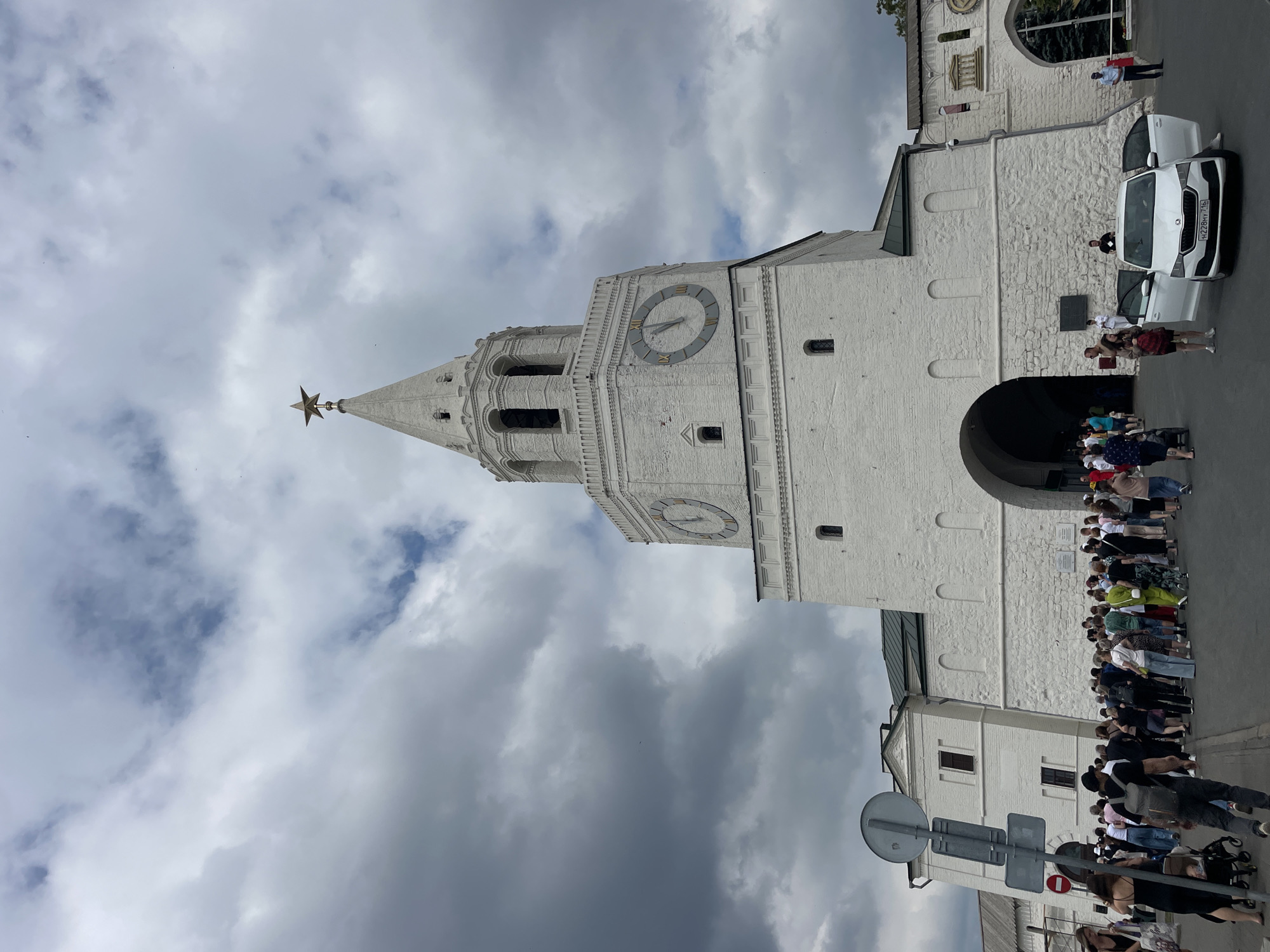 This is a ground-level, eye-level, full-body, exterior shot of the Kul Sharif Mosque in Kazan, Russia, taken on a cloudy day. The mosque features white brick walls, arched entrances, and a prominent white tower topped with a golden star. A large clock face with Roman numerals is visible on the tower. A crowd of people are gathered in front of the mosque, some walking, others standing. A white car with its doors open is parked to the right of the entrance. The sky is filled with dramatic, dark clouds.