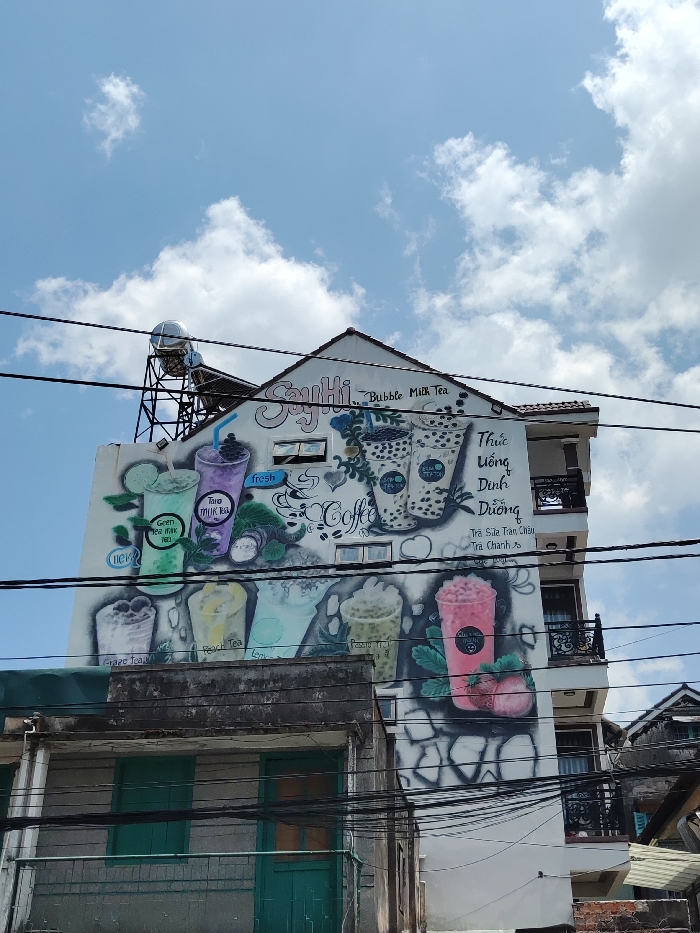 This image captures an urban scene in Thành phố Đà Lạt, Vietnam, primarily featuring the side of a multi-story building. The building has a white facade and a reddish-brown tiled gable roof. A large, colorful mural covers a significant portion of the white wall, advertising beverages. The mural depicts various drinks, including cups labeled "Green Tea Milk Tea," "Taro Milk Tea," "Grape Tea," "Peach Tea," "Longan Tea," and "Passion Tea." Several illustrations of bubble milk tea with dark pearls are visible, alongside a pink blended drink with strawberries. Text on the mural includes "Say Hi... Bubble Milk Tea," "fresh," "Coffee," and Vietnamese phrases such as "Thức Uống Dinh Dưỡng" (Nutritious Drinks), "Trà Sữa Trân Châu" (Bubble Milk Tea), and "Trà Chanh" (Lemon Tea).
A metallic cylindrical water tank, supported by a black metal frame, is mounted on the roof of the building, positioned to the left of the gable. Multiple dark electrical or communication wires run horizontally across the foreground, partially obscuring the building. On the right side of the main building, two balconies with black wrought-iron railings are visible. Below the main building, to the left, is an older, lower structure with a grey exterior and two closed green wooden doors or shutters. A section of another building with a dark roof is partially visible on the far right. The sky above is bright blue with scattered white cumulus clouds, indicating daylight conditions. No people are visible in the scene.