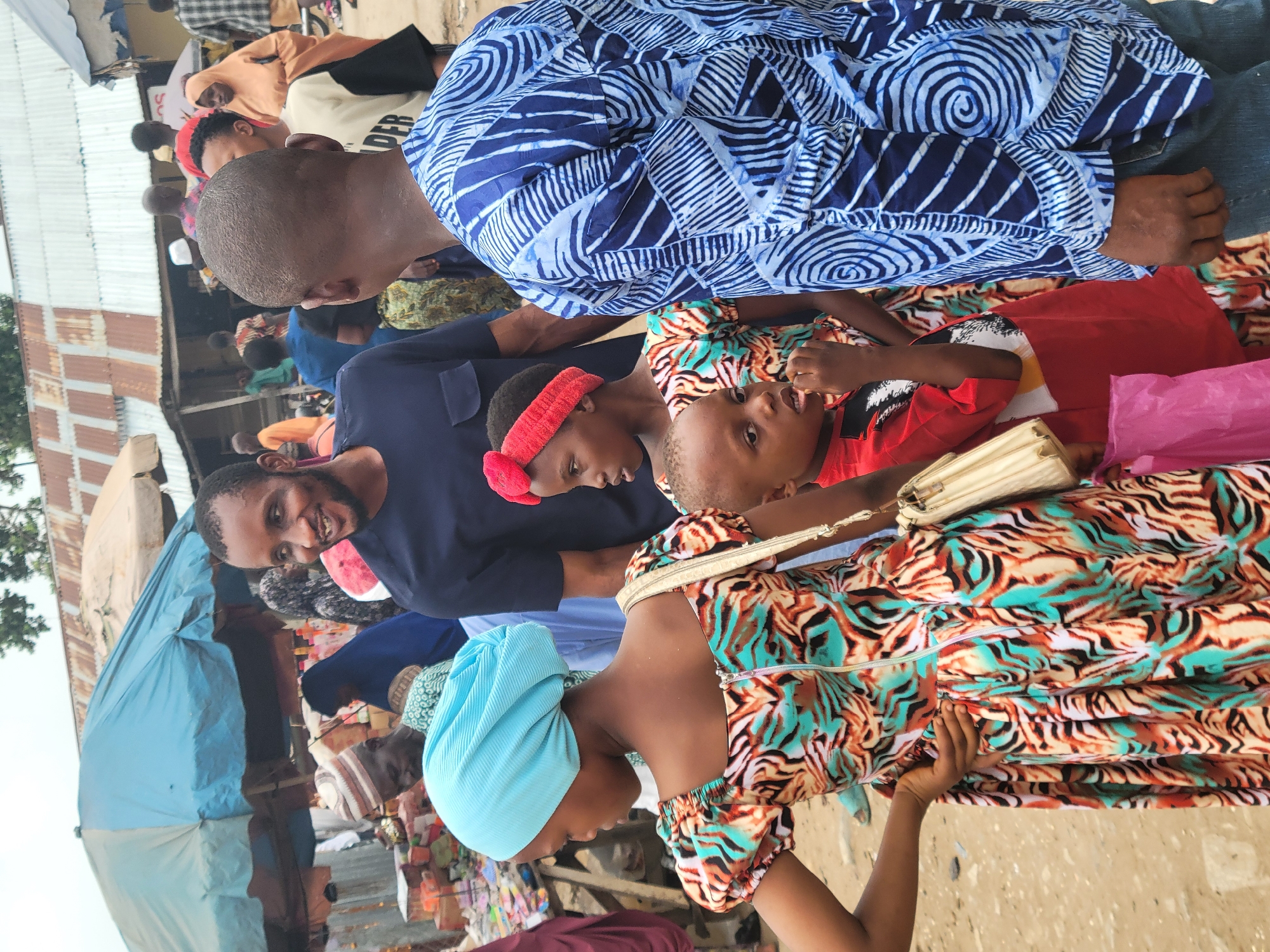 The image depicts a crowded scene, possibly a marketplace in Madalla, Nigeria. People of various ages are present. A woman with a light blue head covering and a colorful patterned dress stands near the lower edge of the frame. A man in a dark blue shirt is smiling and looking at the camera. Two children, one wearing a red headband and the other in a red shirt, are in the scene. Another man is wearing a blue patterned shirt. A blue tent-like structure is visible in the background, along with various stalls and other individuals.